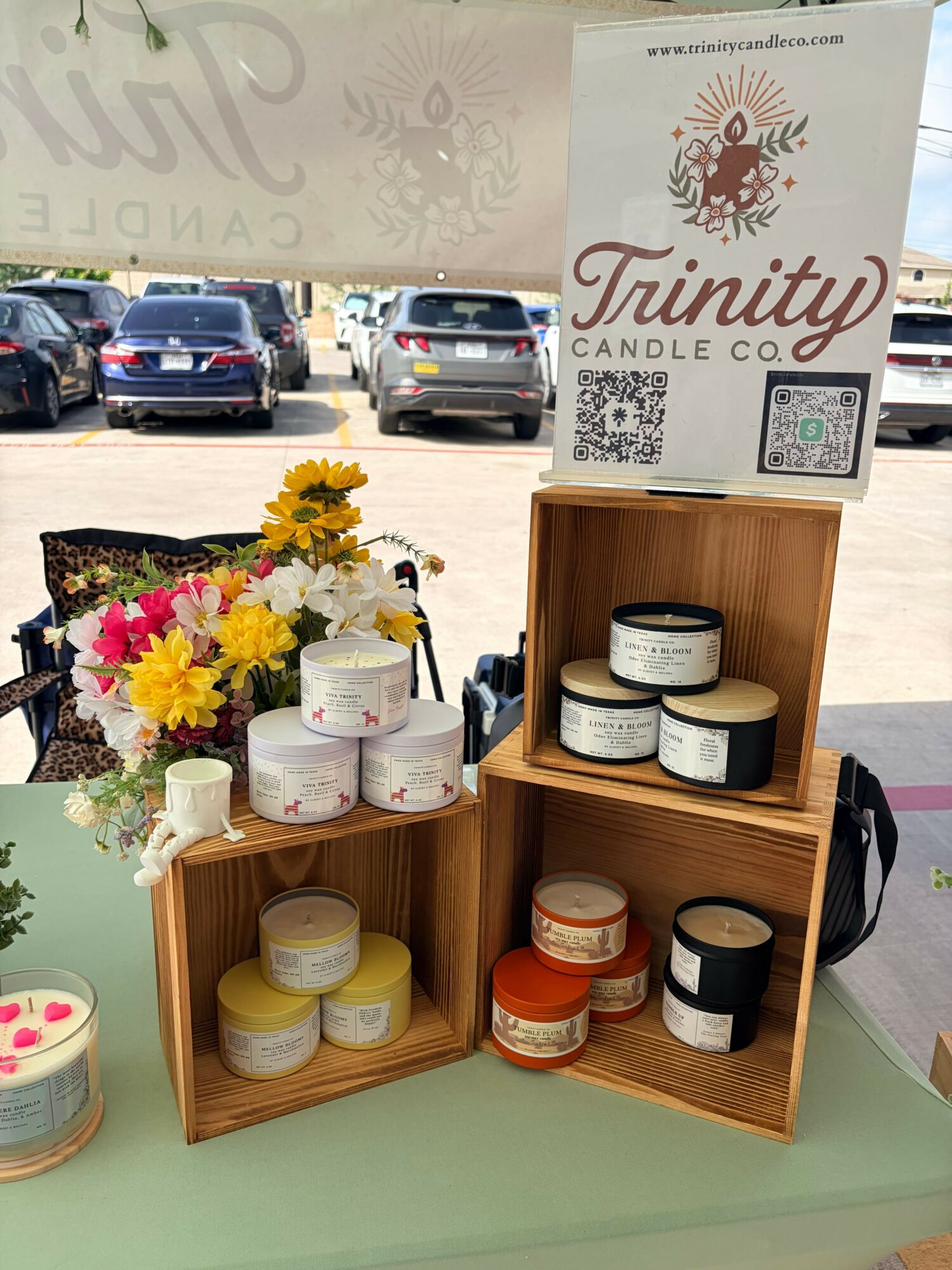 Display of candles on wooden shelves with flowers, in a store setting, with cars visible outside through a window.