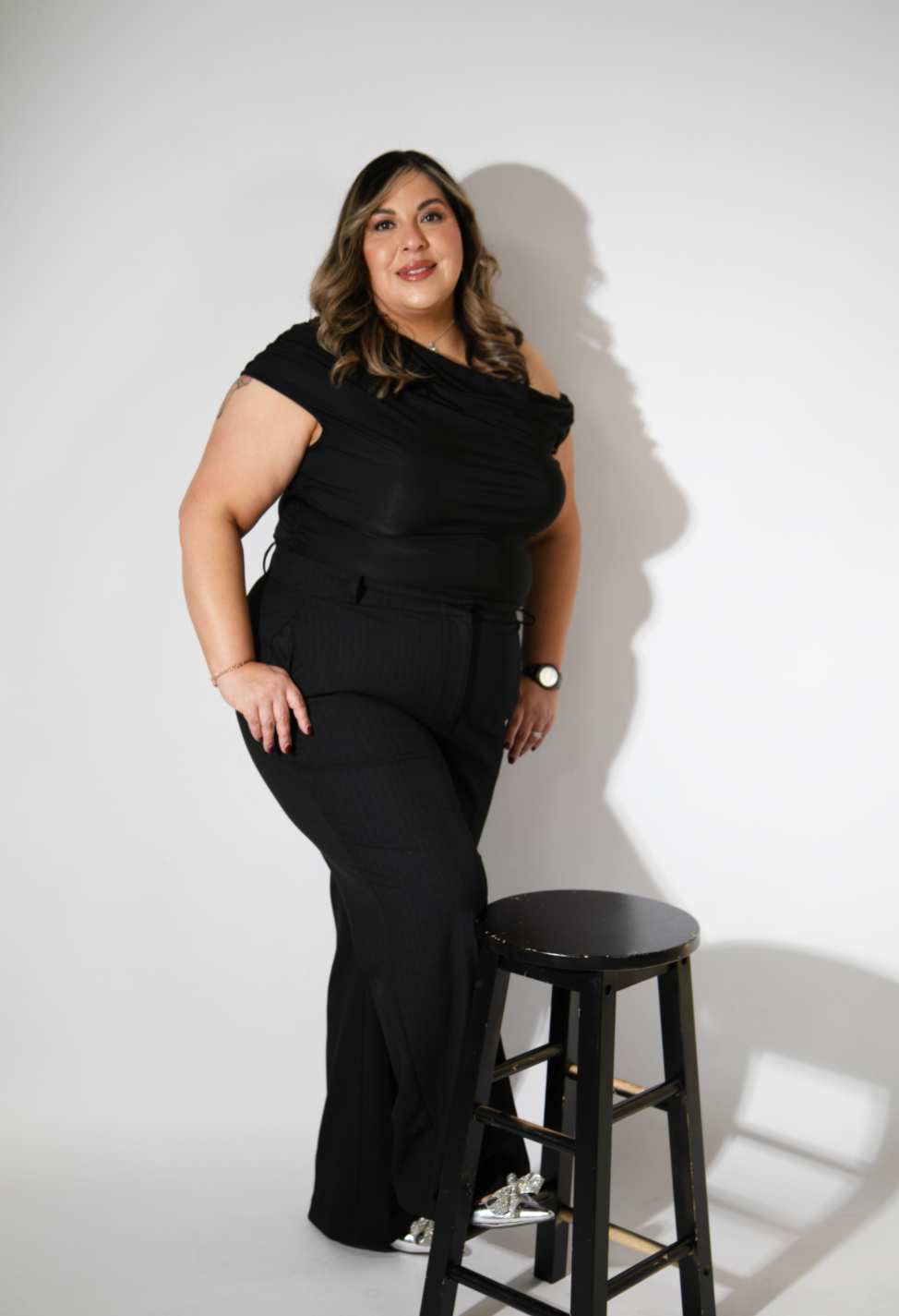 Woman in black outfit standing next to a stool, smiling, with a plain white background.