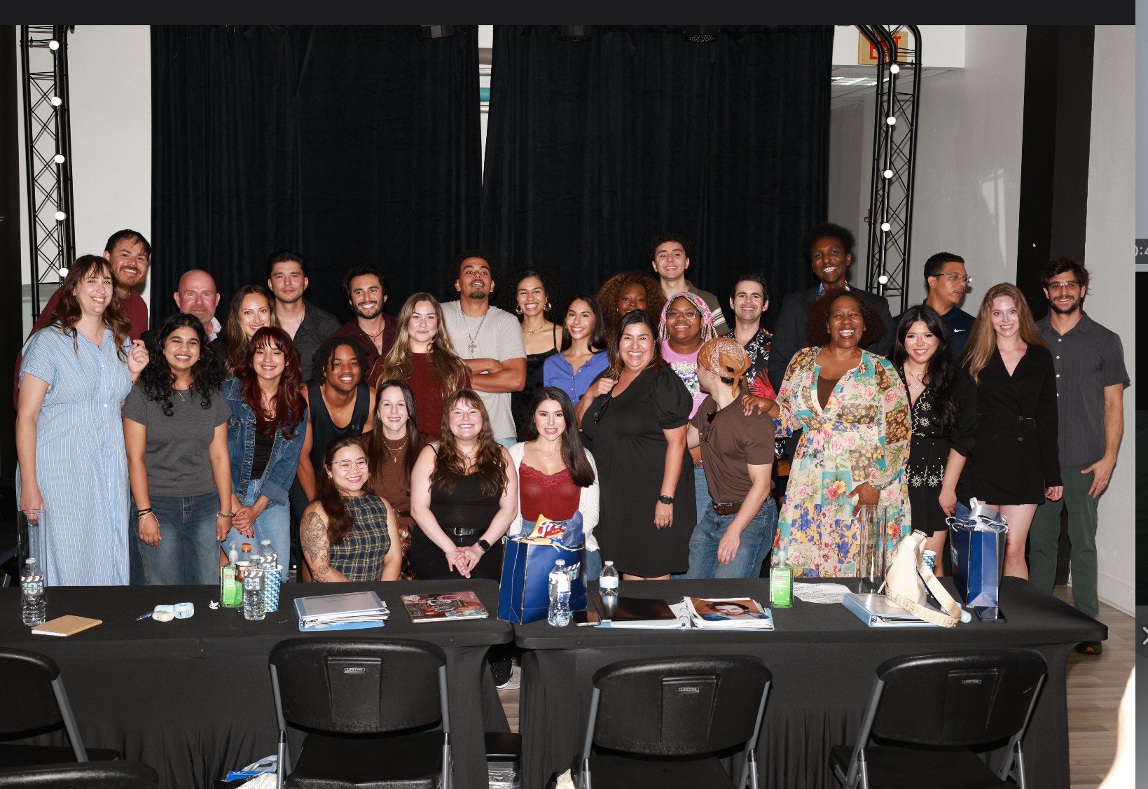 Group of people posing for a photo behind a table with chairs, some holding drinks, in a room with black curtains.