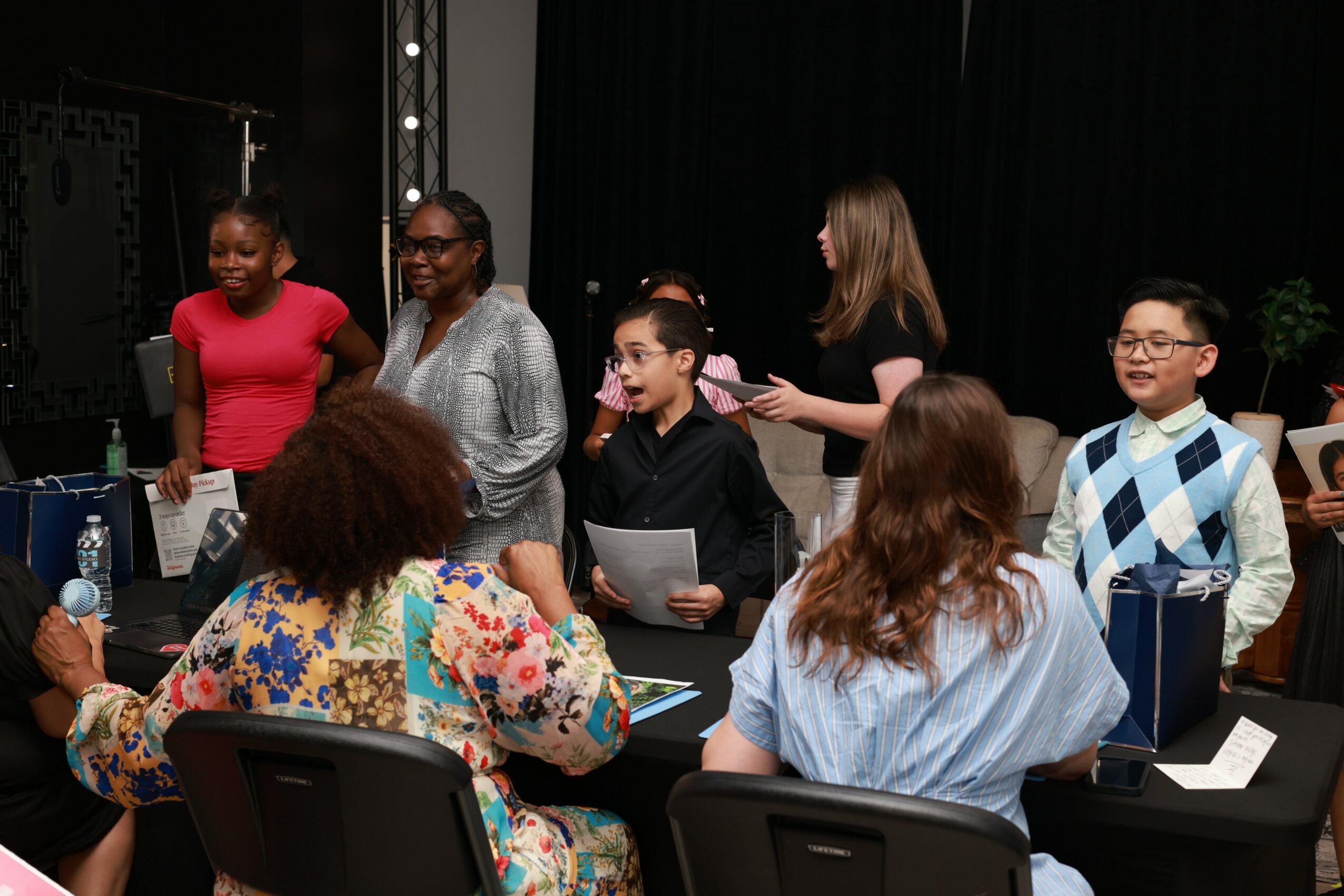 Group of people gathered around a table, some holding papers, in a room with a black curtain background.