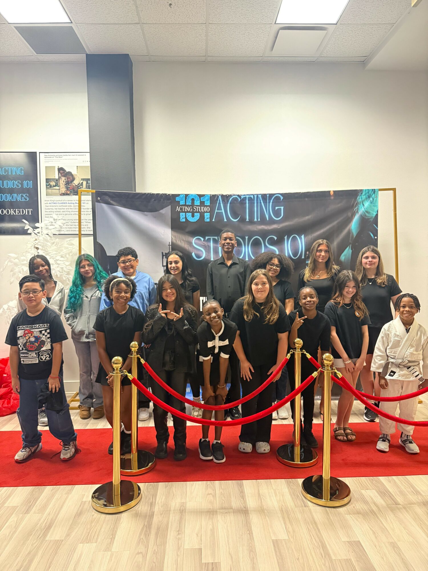 Group of people standing behind a red velvet rope barrier in front of a banner with text, indoors with wooden flooring.