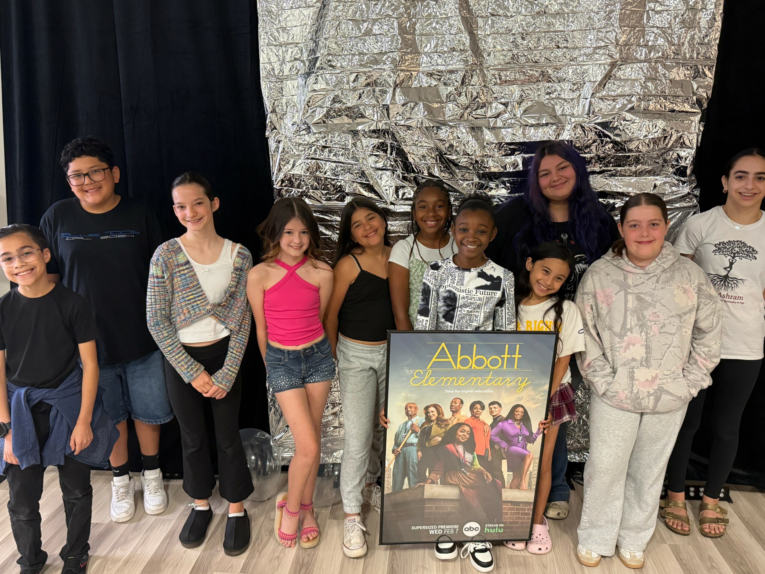 Group of children and an adult holding a poster, standing in front of a silver backdrop, smiling.
