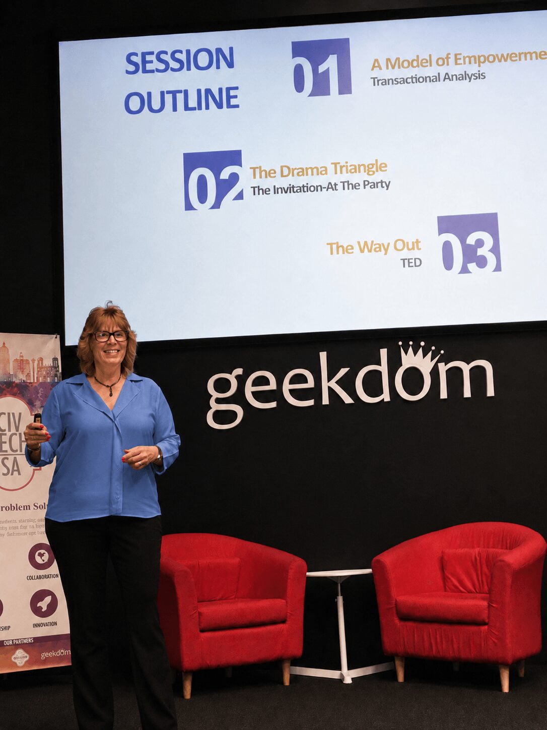 Woman standing on stage with a presentation slide behind her, two red chairs in front, and a black backdrop with 'geekdome' logo.