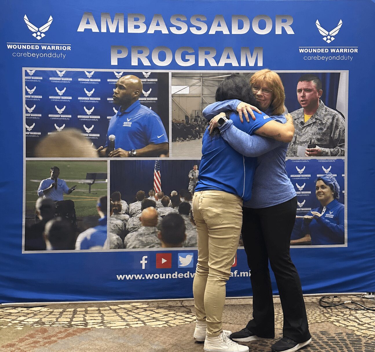 Two women hug in front of a blue banner with images of people and text, one woman has red hair, the other has dark hair.