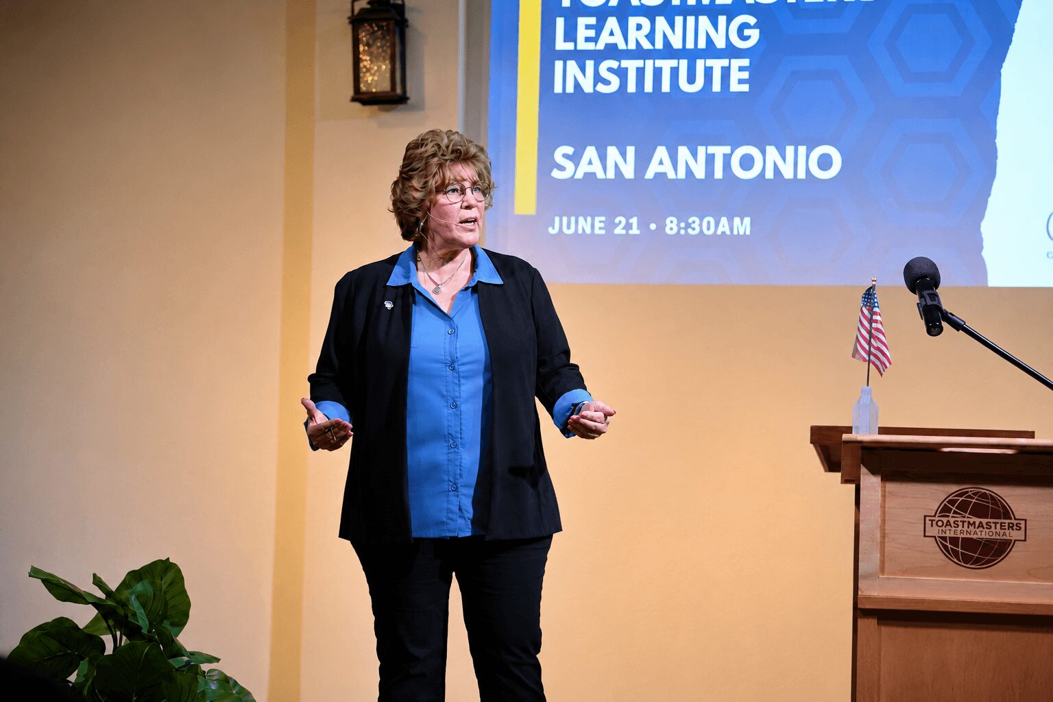 Woman with curly hair and glasses speaking in front of a screen, holding a remote, with a podium nearby, in a room with a plant.