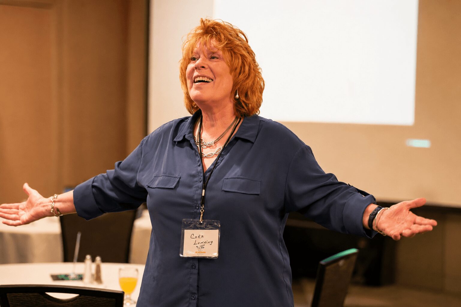 Woman with red hair smiling and gesturing with arms open in a conference room.