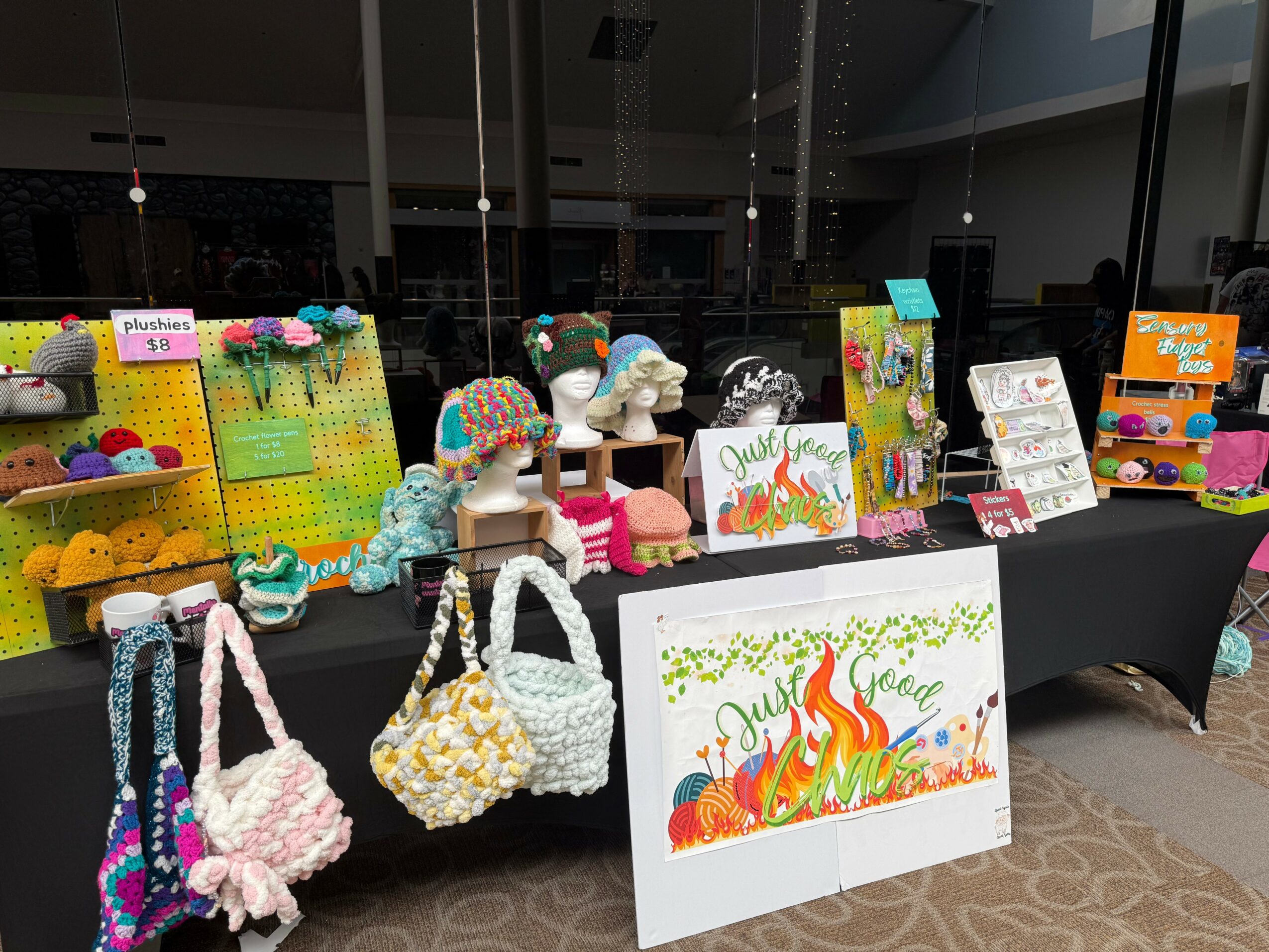 Display table with colorful handmade crochet items, signs, and artwork at an indoor craft fair.