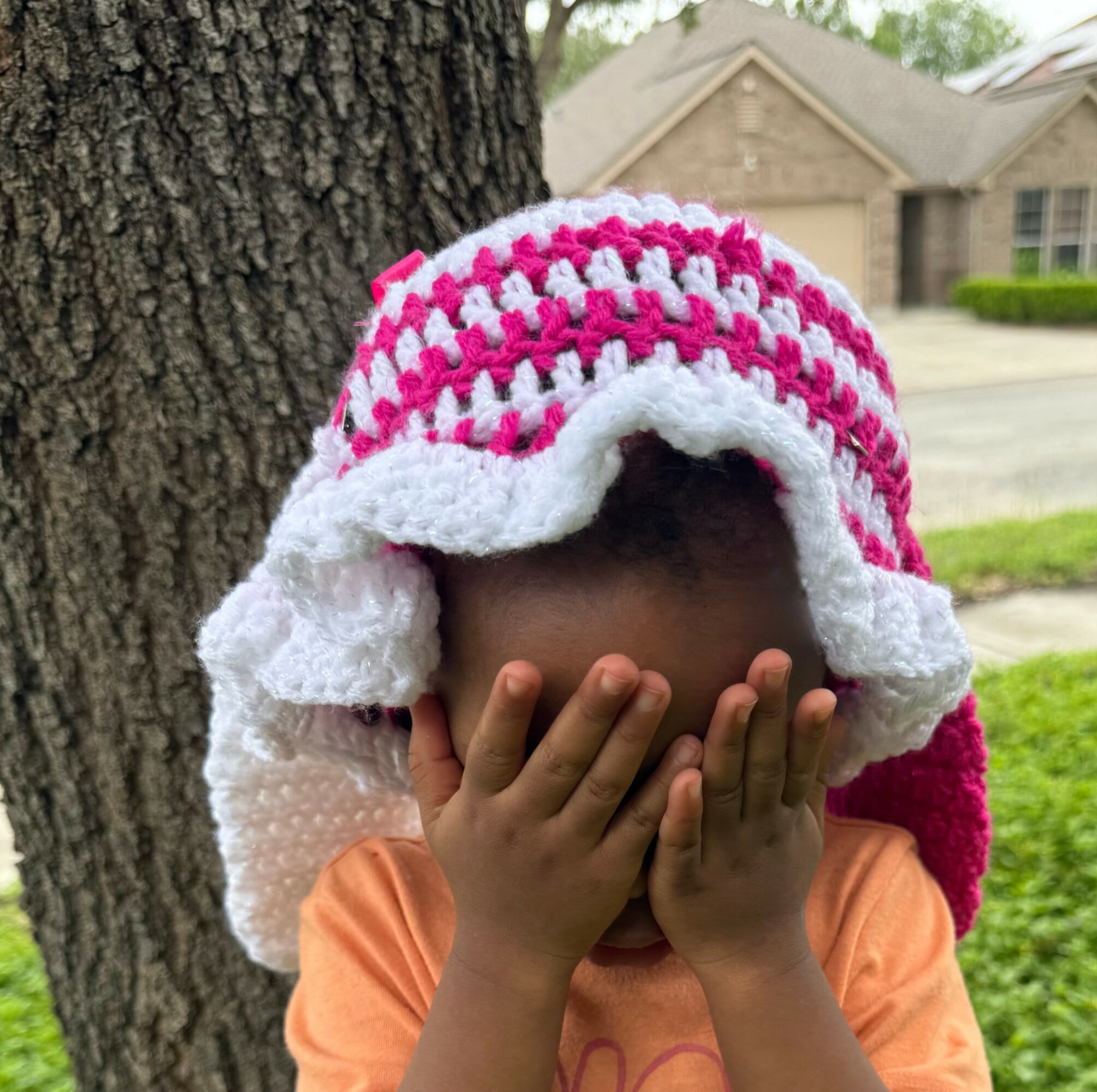 Child covering face with hands, wearing a pink and white crocheted hat, standing outdoors near a tree.