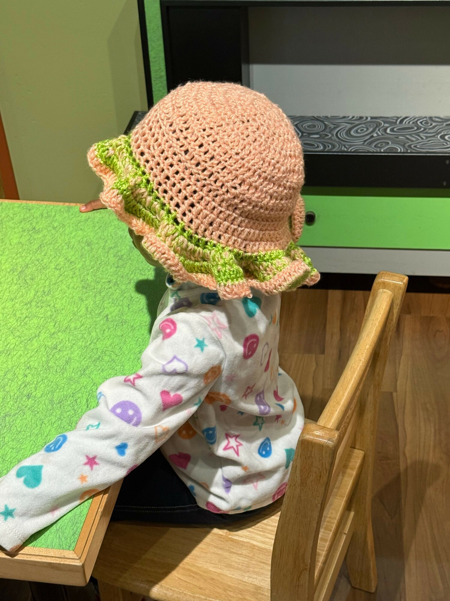 Child wearing a pink hat and colorful pajamas, sitting on a chair with a wooden floor and green wall in background.