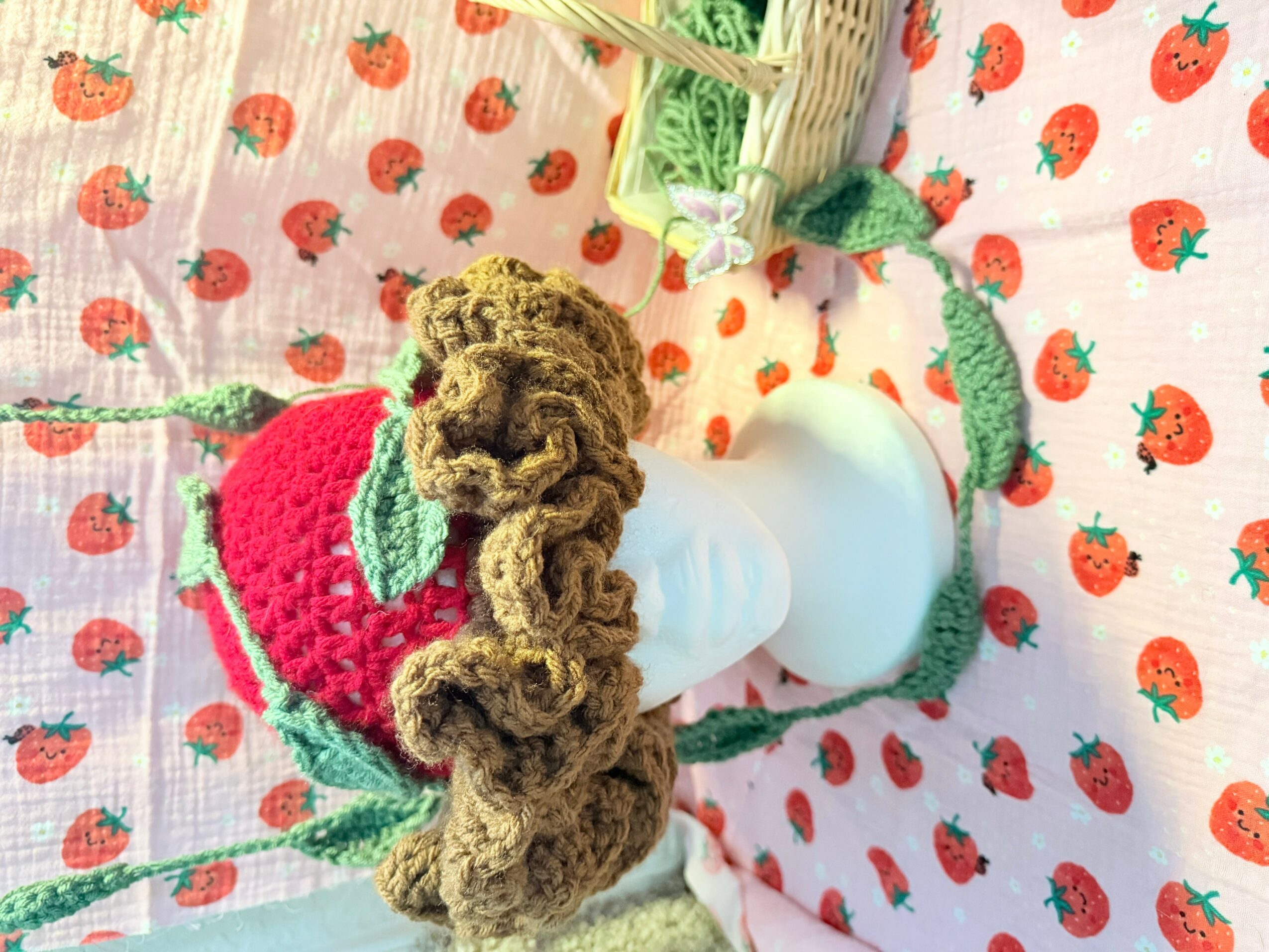 A plush strawberry with a brown curly tail and green leaves, placed on a strawberry-patterned fabric background.