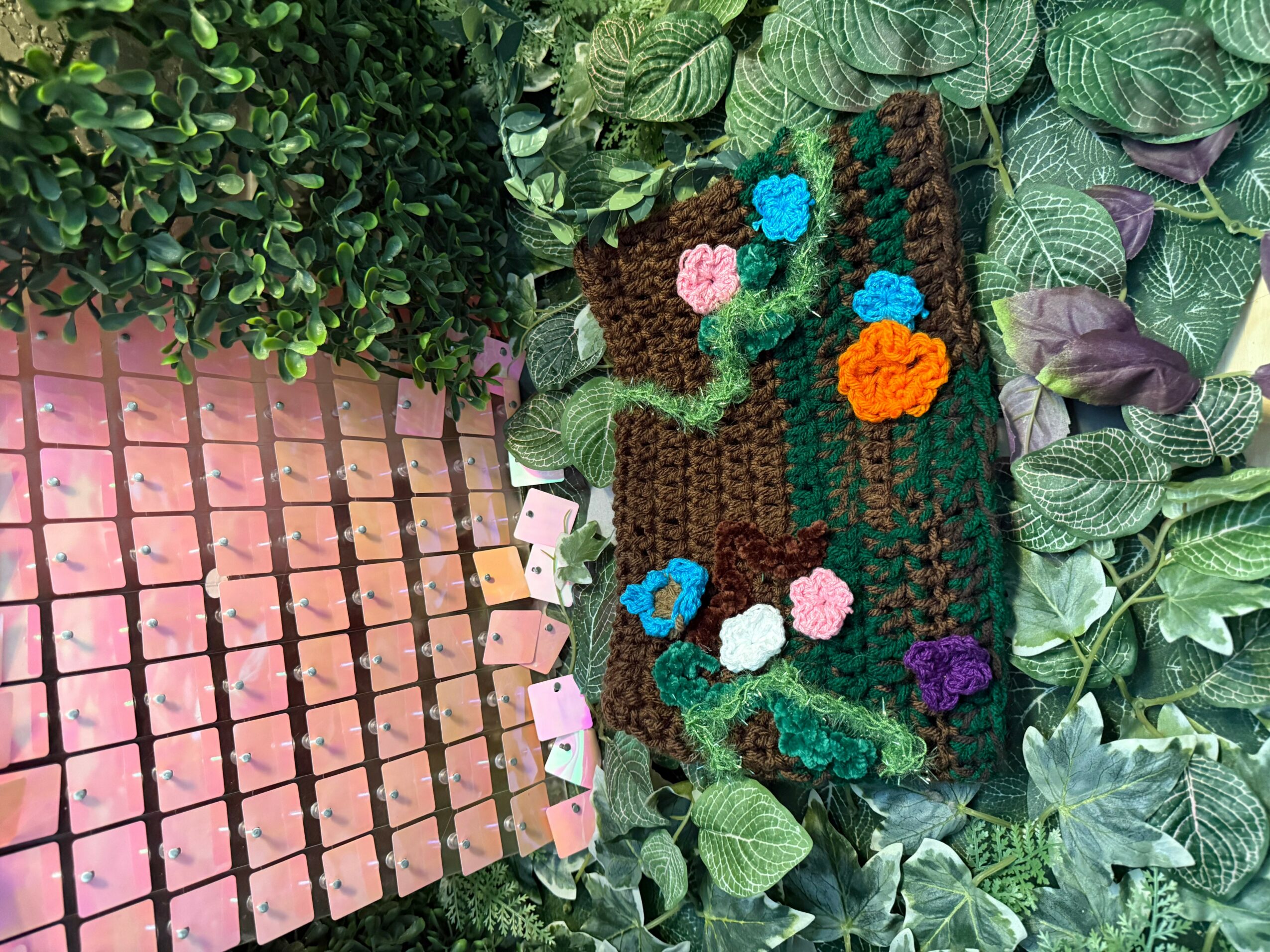 Colorful knitted bag with flowers on green leaves, surrounded by various green plants and a pink tiled surface.