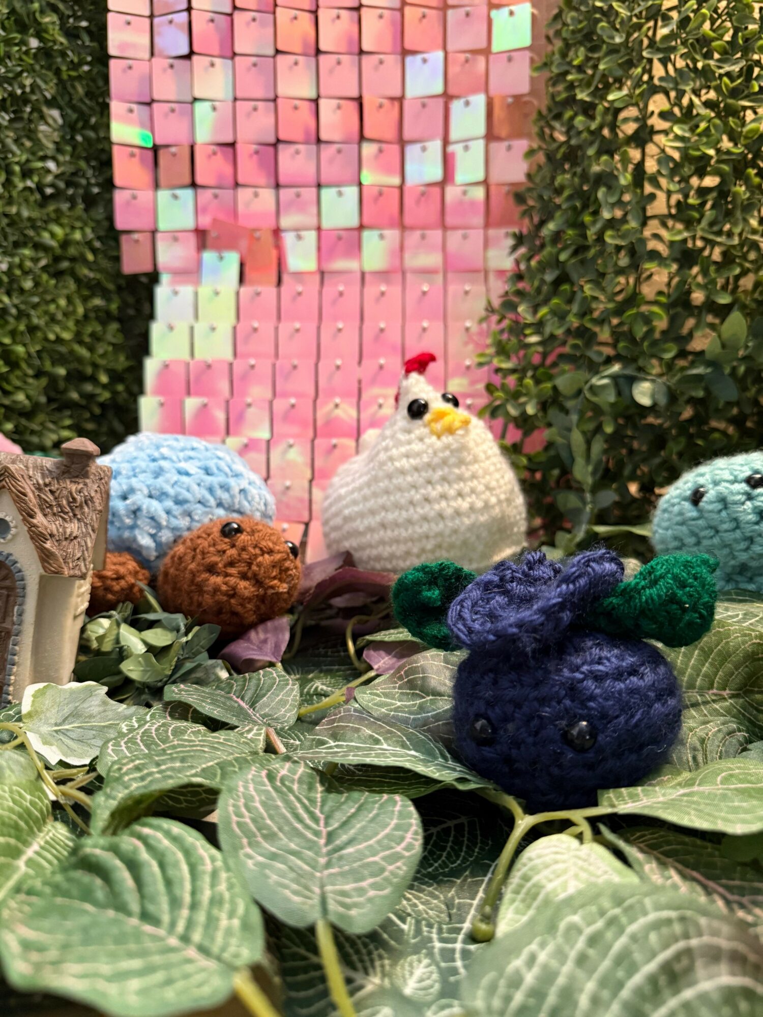 Colorful knitted animal toys among green leafy plants and a pink sticky note wall.