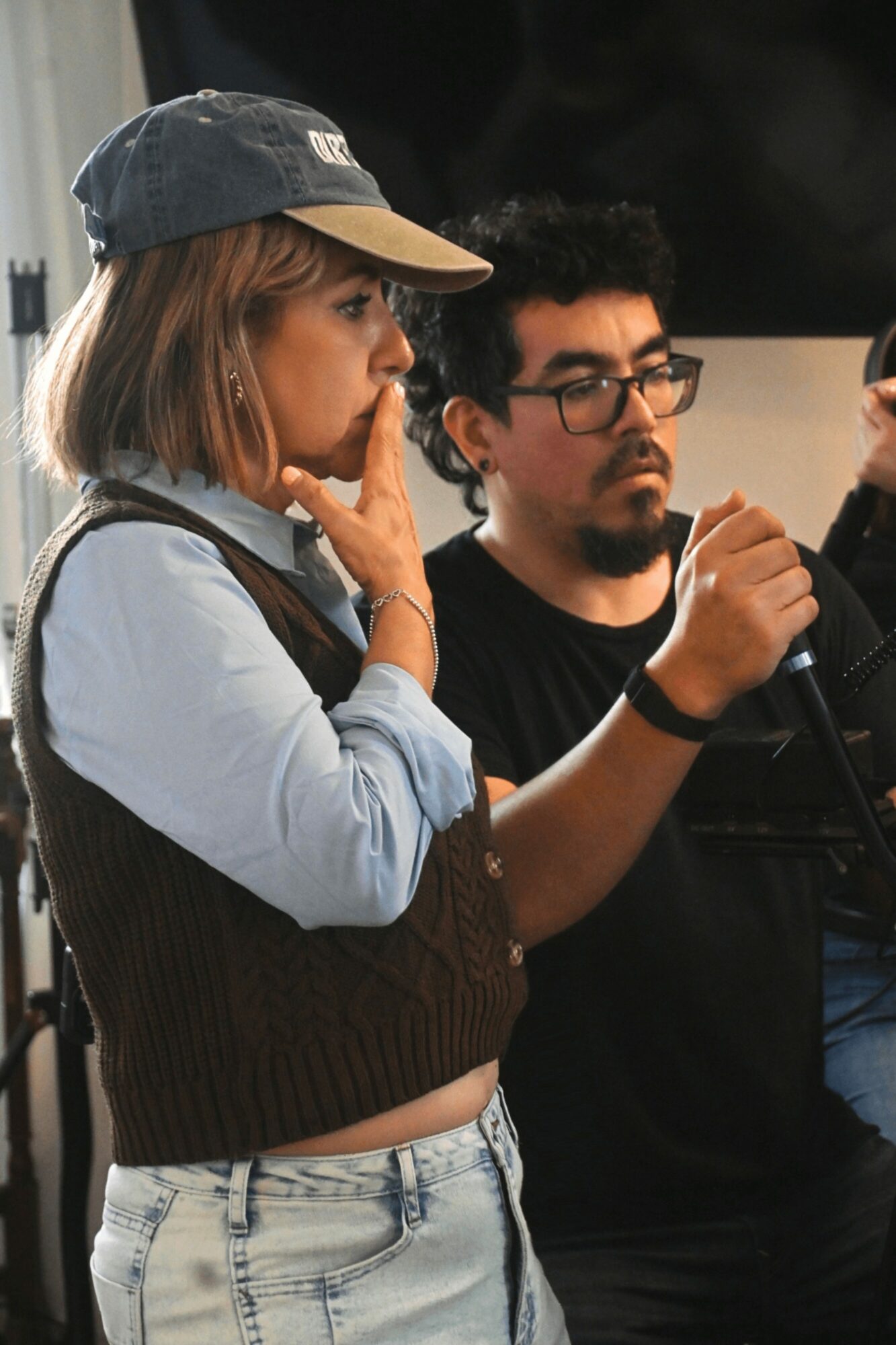 Two people, a woman and a man, are engaged in a conversation or activity indoors. The woman has a cap and is touching her face, the man is holding an object and wearing glasses.