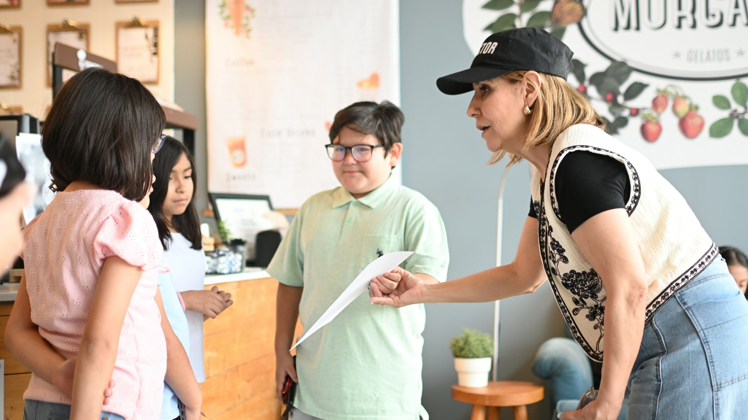 Woman handing paper to children in a store or cafe, with a counter and decorative wall in background.