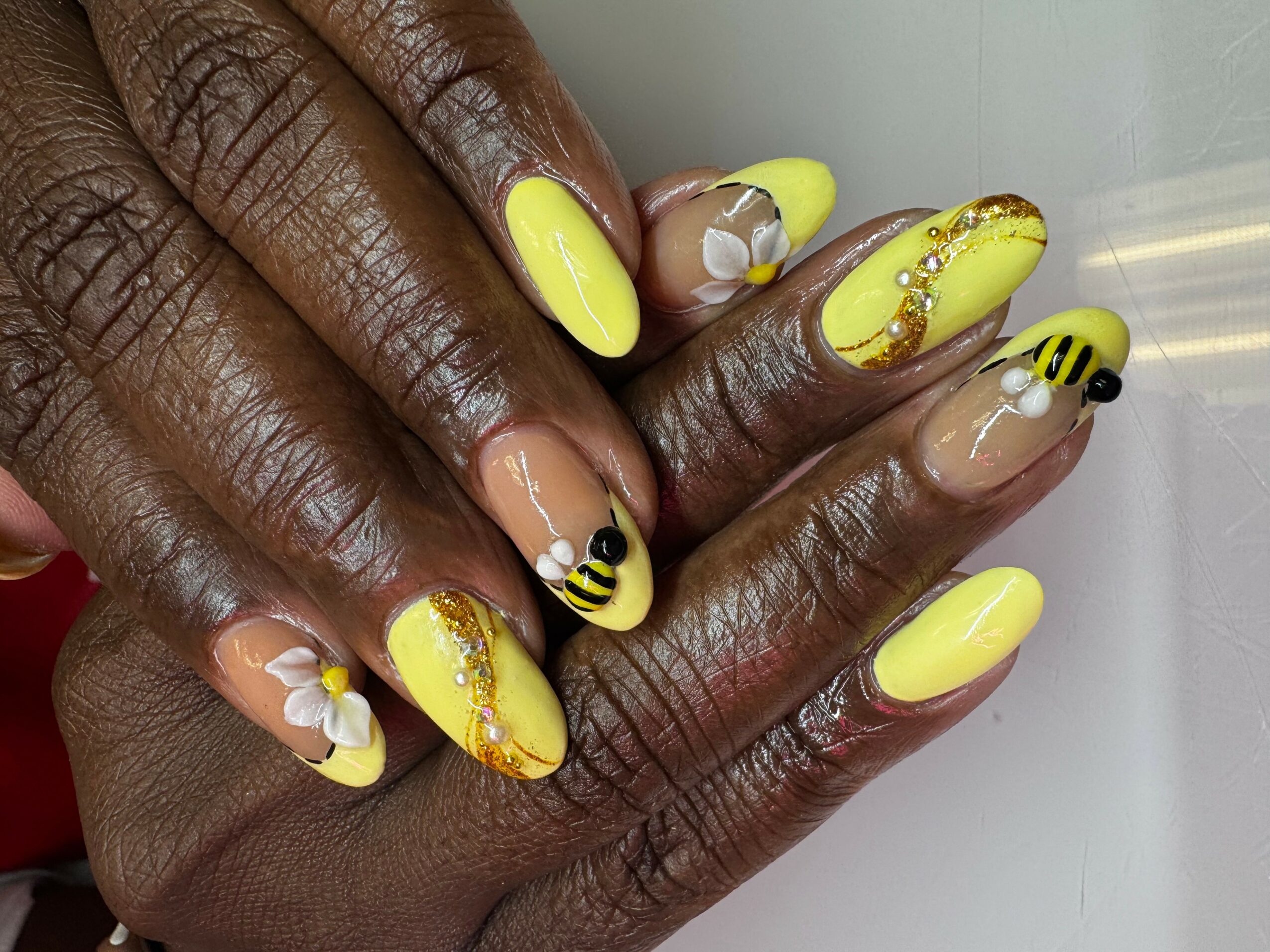 Close-up of hands with yellow, bee-themed and floral nail art, including yellow, black, and white designs, and rhinestones.