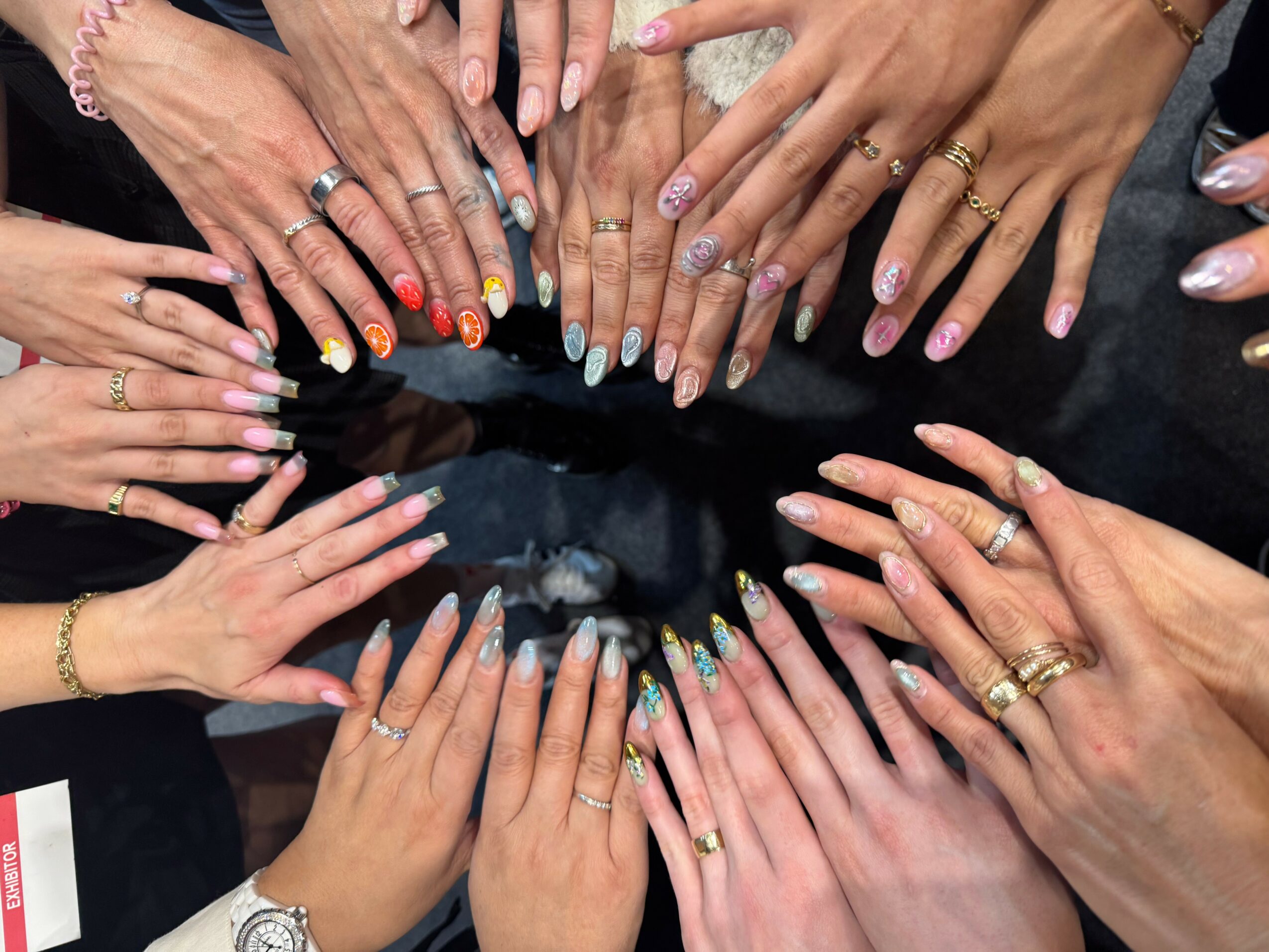 Multiple hands with various rings and nail polish arranged in a circle, showing diverse skin tones and manicures.