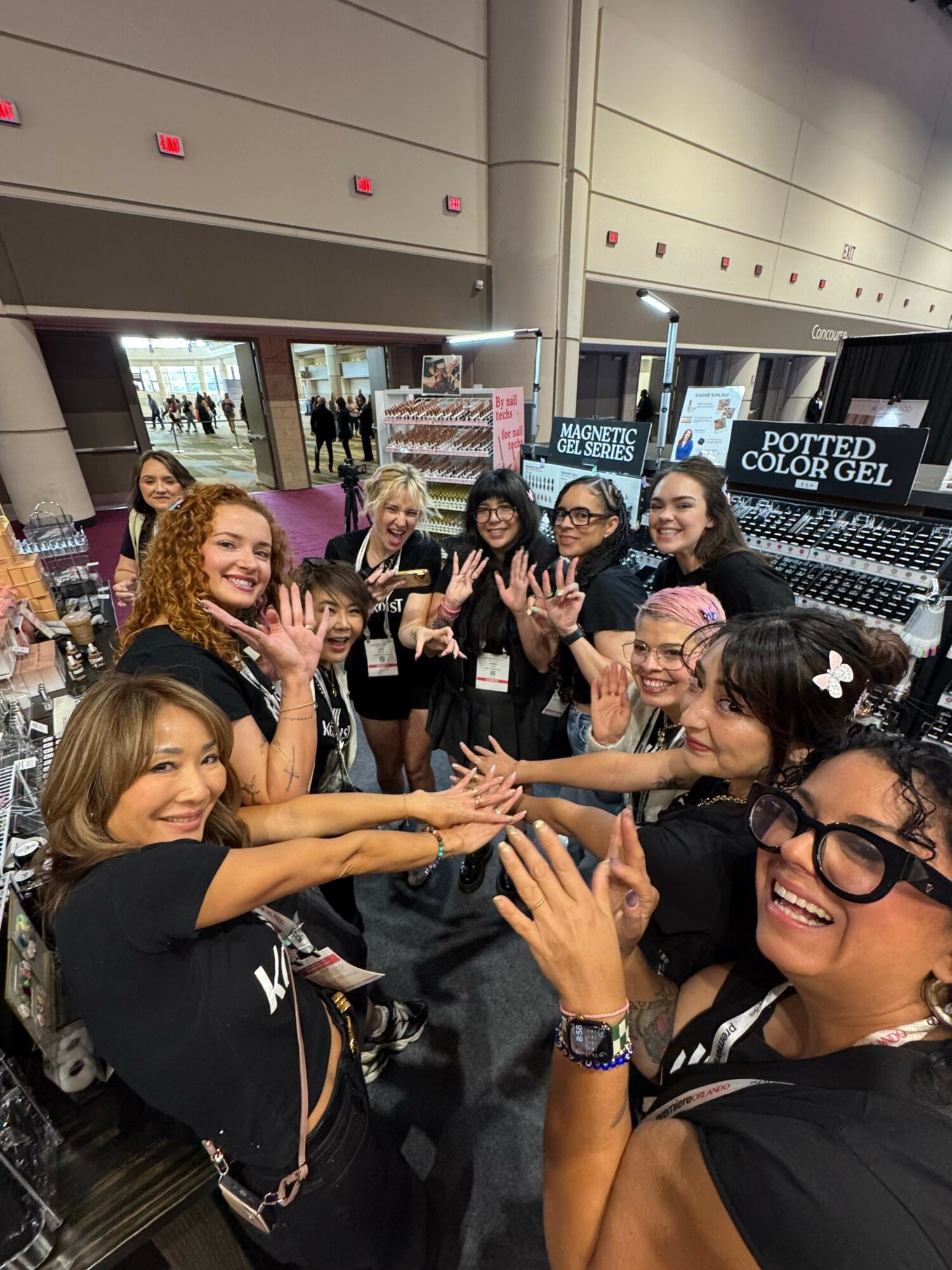 Group of women smiling and making hand gestures at an indoor event, some wearing glasses and casual clothing.