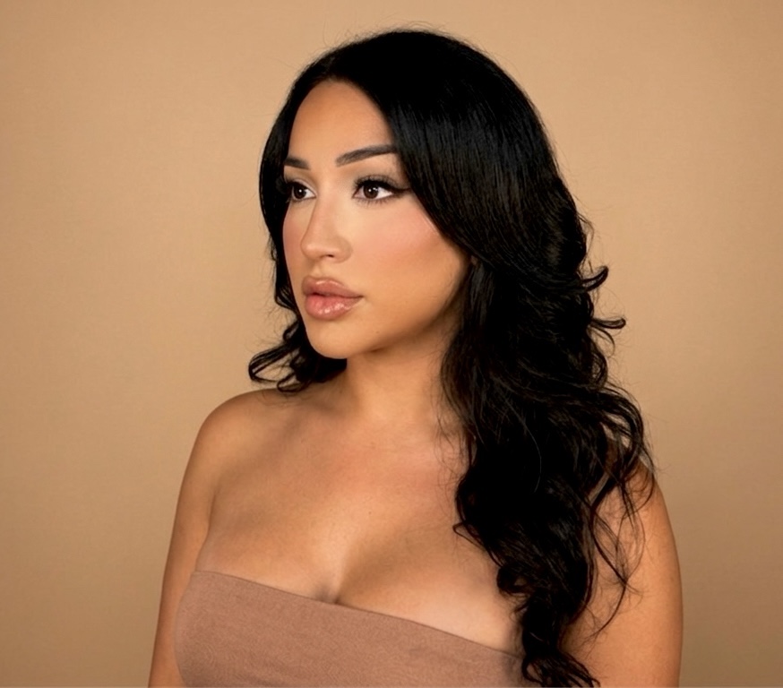 Woman with long black hair and makeup, wearing a strapless top, against a beige background.
