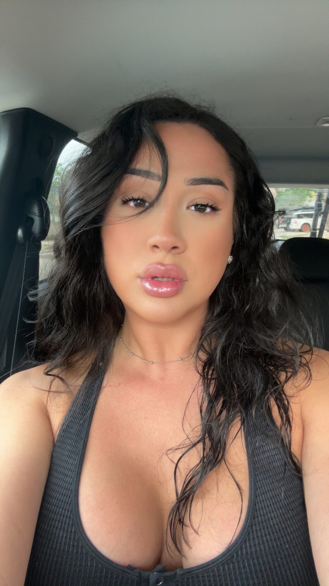 Young woman with wavy dark hair and makeup, wearing a black top, sitting in a car, looking at the camera.