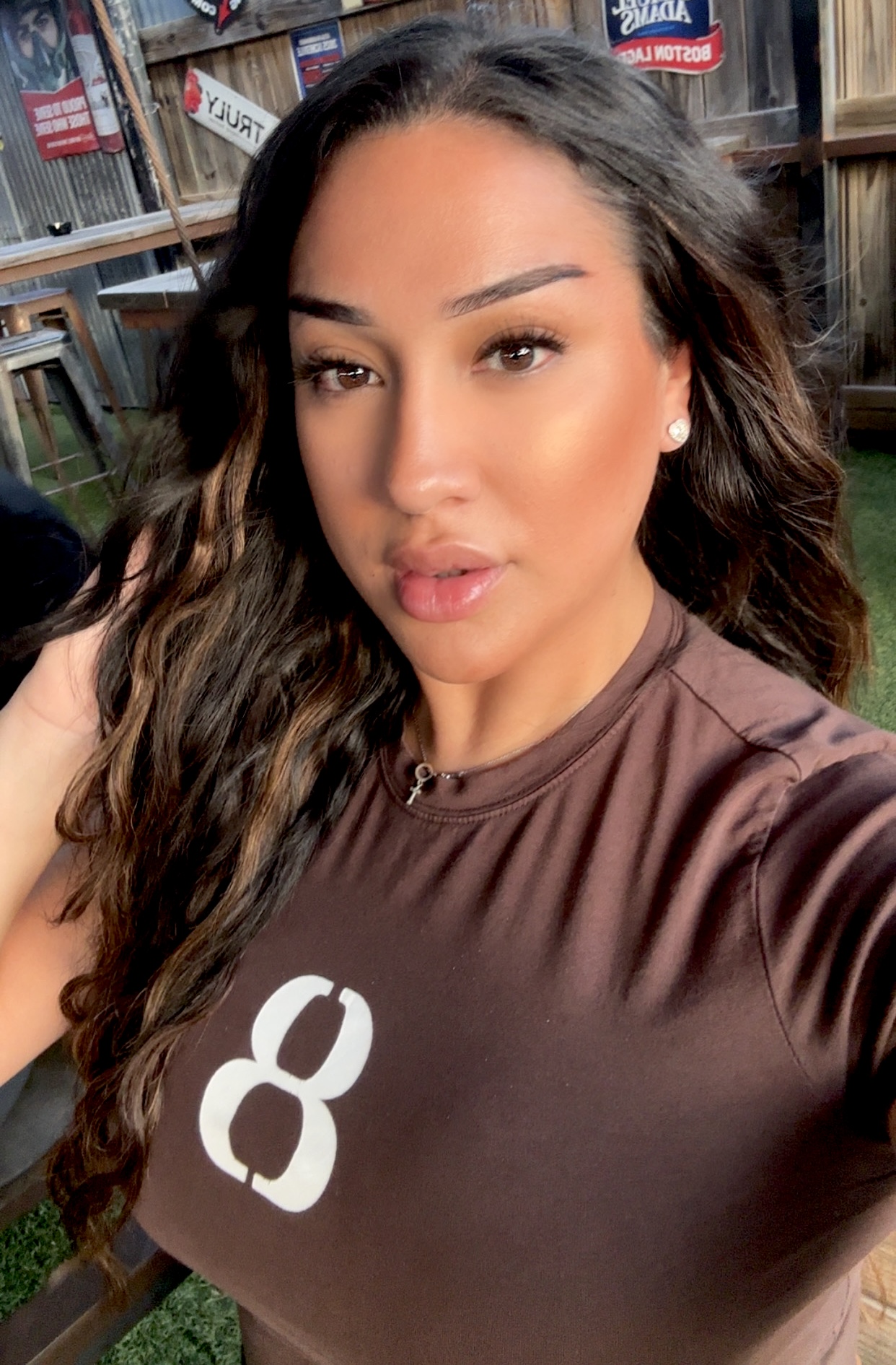 Young woman with long wavy hair taking a selfie outdoors, wearing a brown shirt with a white number eight, background of wooden structures.