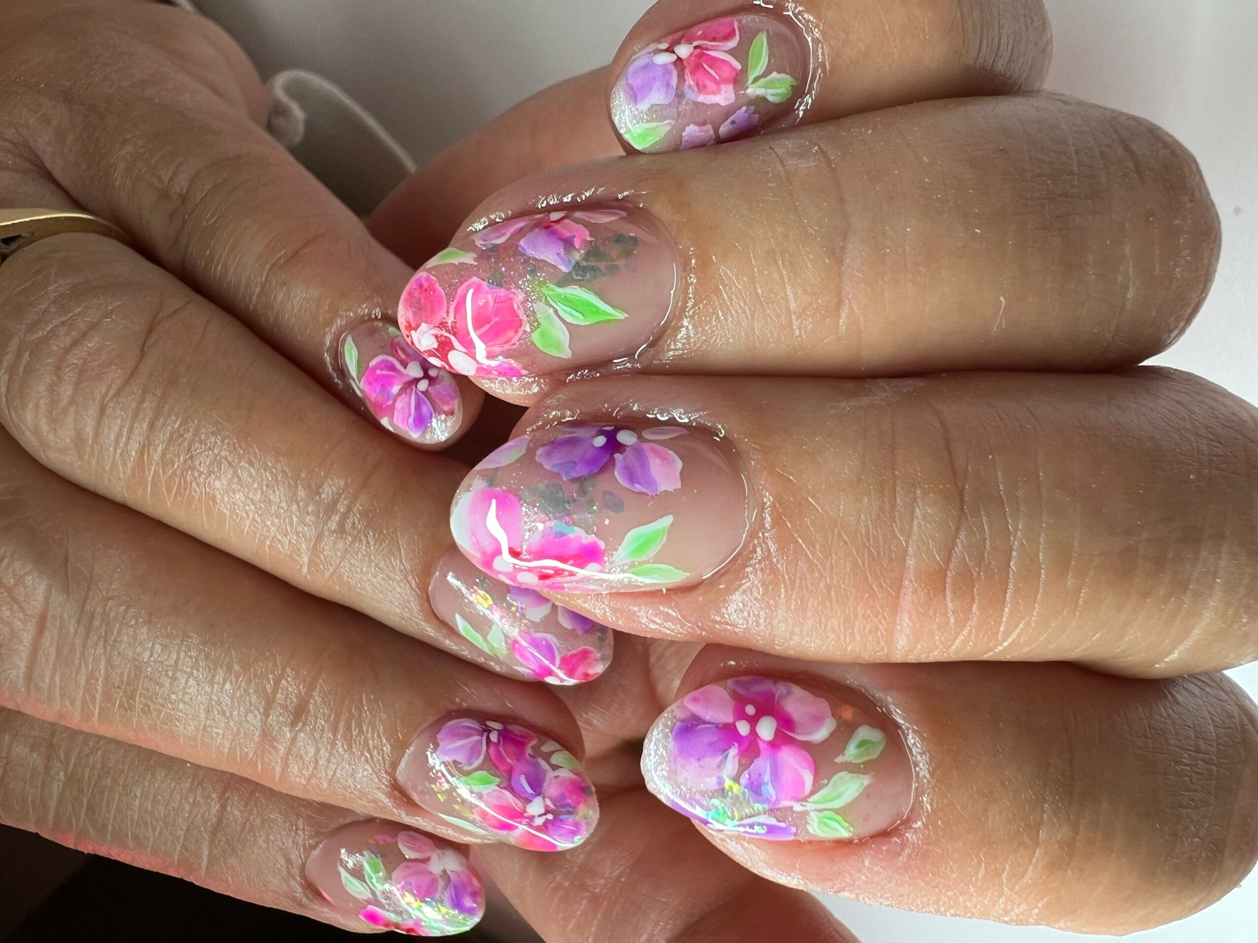Close-up of hands with floral nail art on fingernails, featuring pink, purple, and green flowers on a clear background.