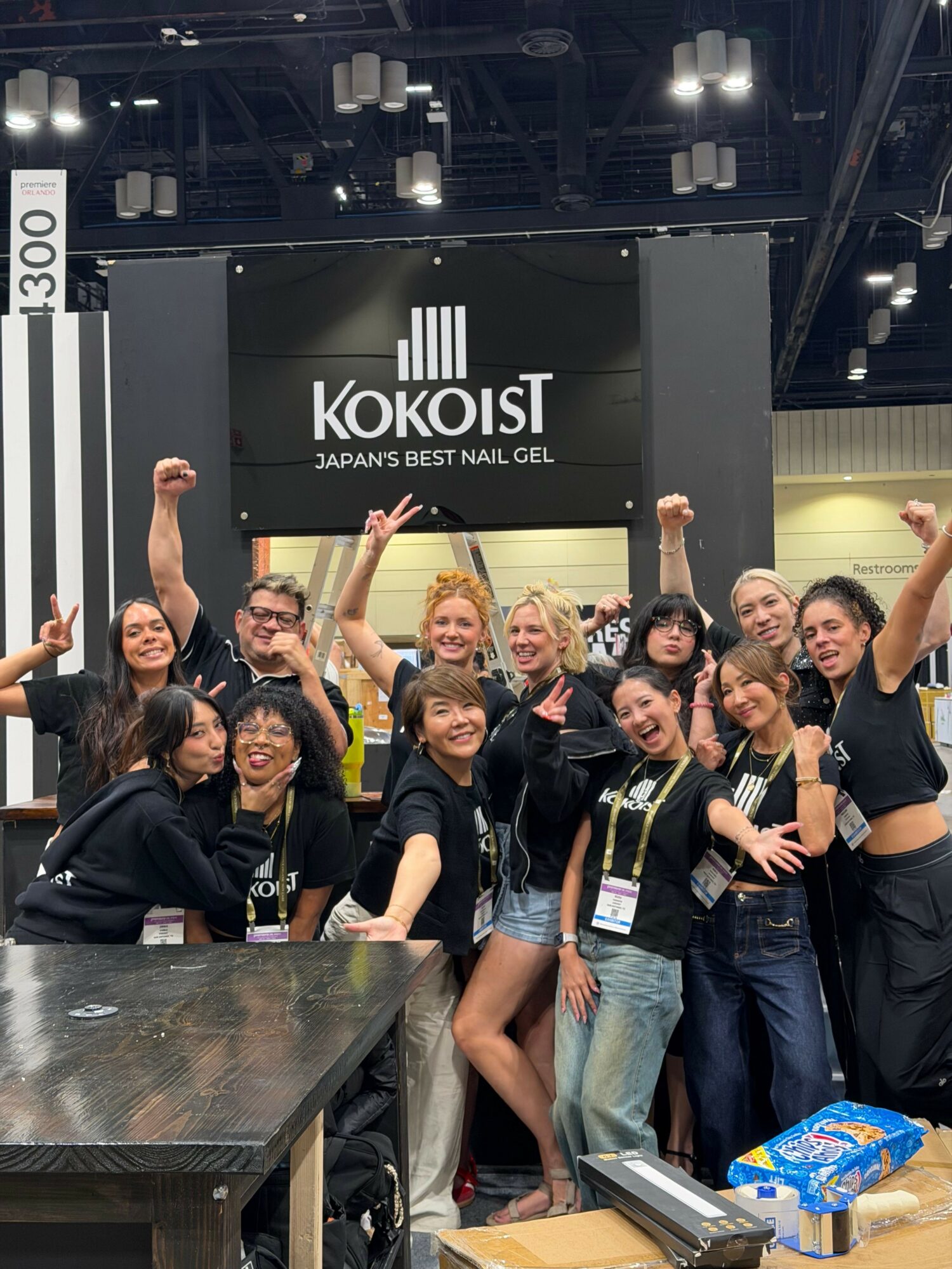 Group of people celebrating at an event, some raising hands, in front of a KoKoIST banner, with a table nearby.