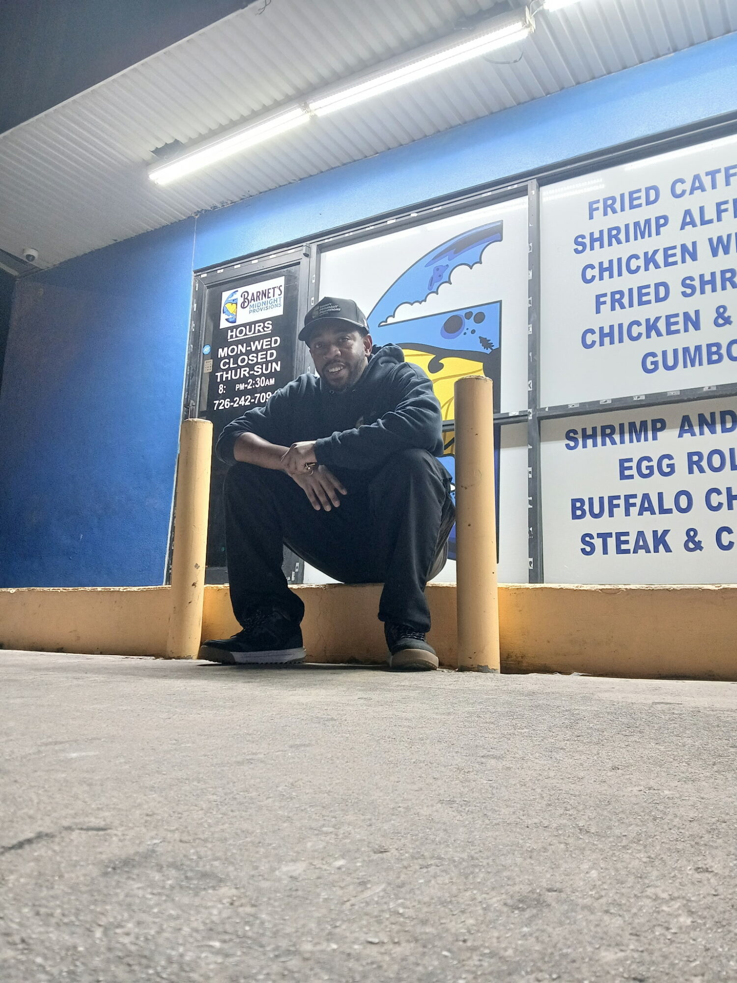 Person sitting on a yellow barrier outside a fast-food restaurant, holding a book, with menu signs in the background.