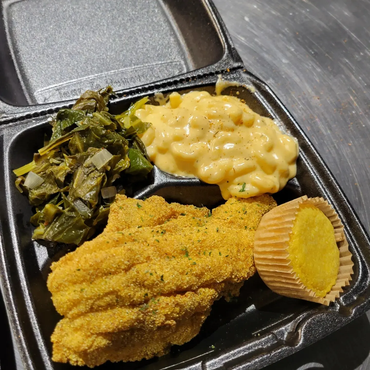 Fried fish fillet with mashed potatoes, green beans, and a cupcake in a black takeout container.
