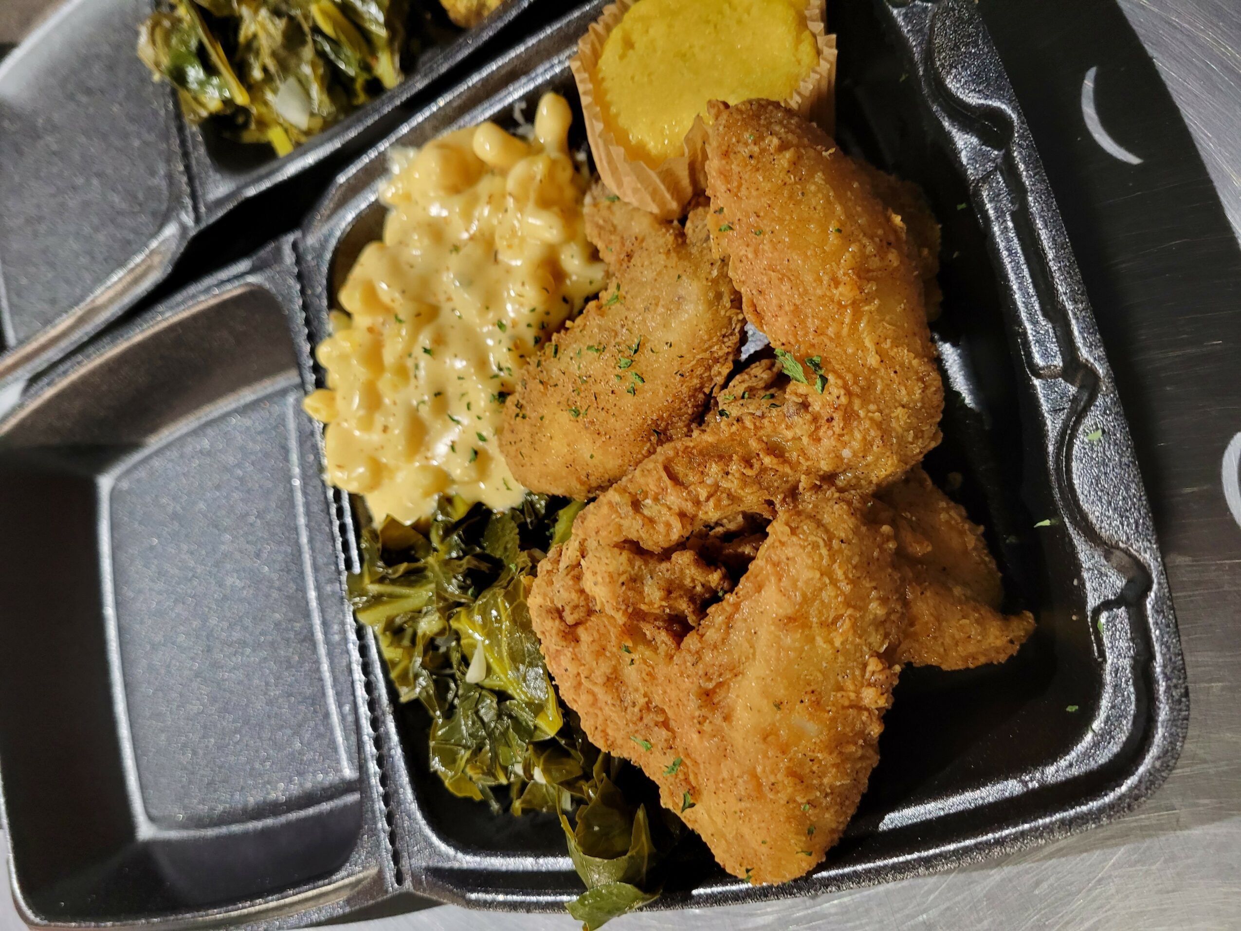 Fried fish fillets with lemon wedge, macaroni and cheese, and greens in a black takeout container.