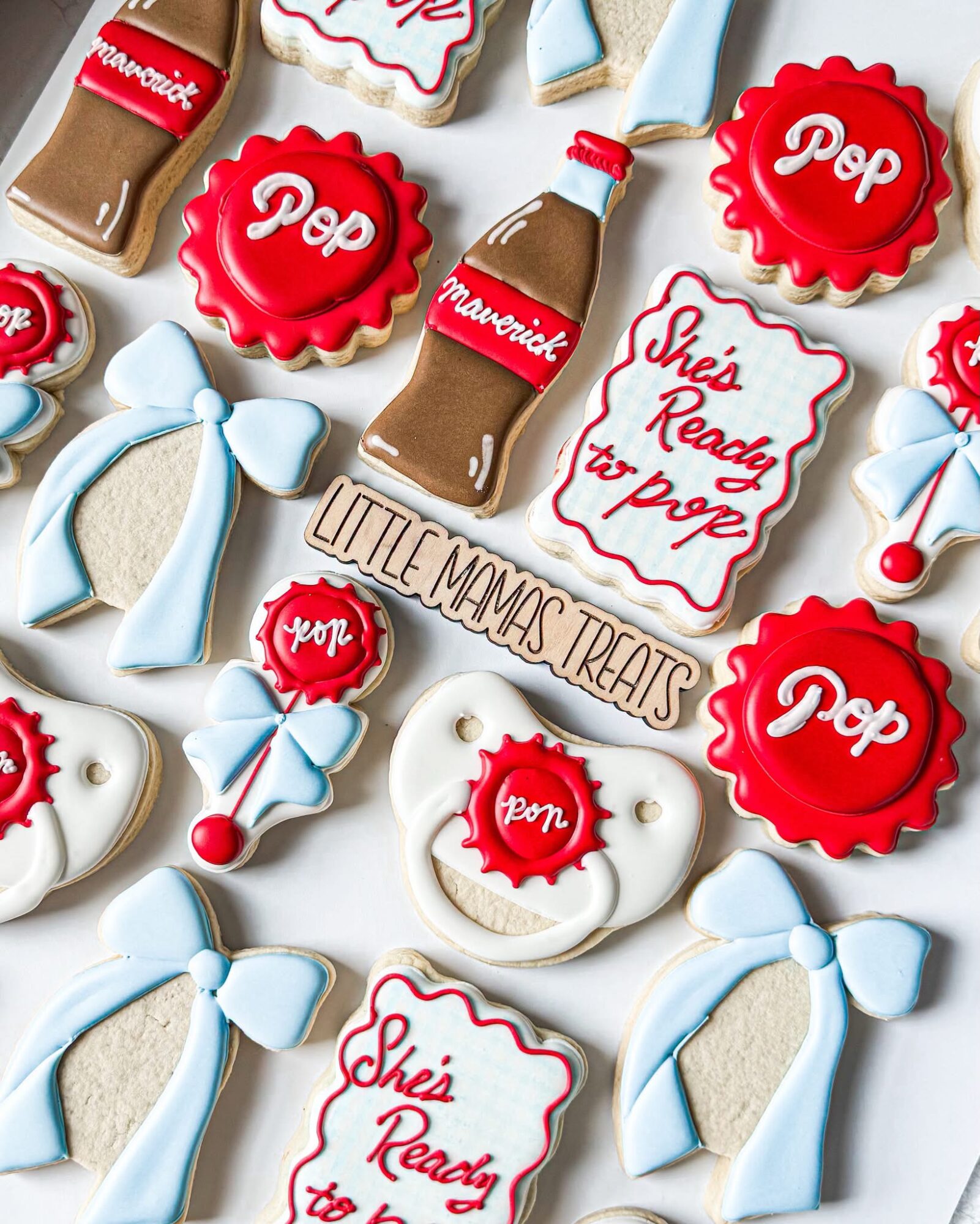 Assorted cookies decorated with pop bottles, bows, and messages for a baby shower, including 'She's Ready to Pop' and 'Little Man's Treats'.