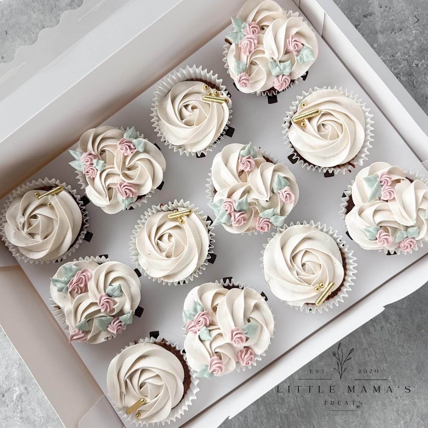 Box of decorated cupcakes with white frosting and pink and blue floral accents, arranged in a white box.