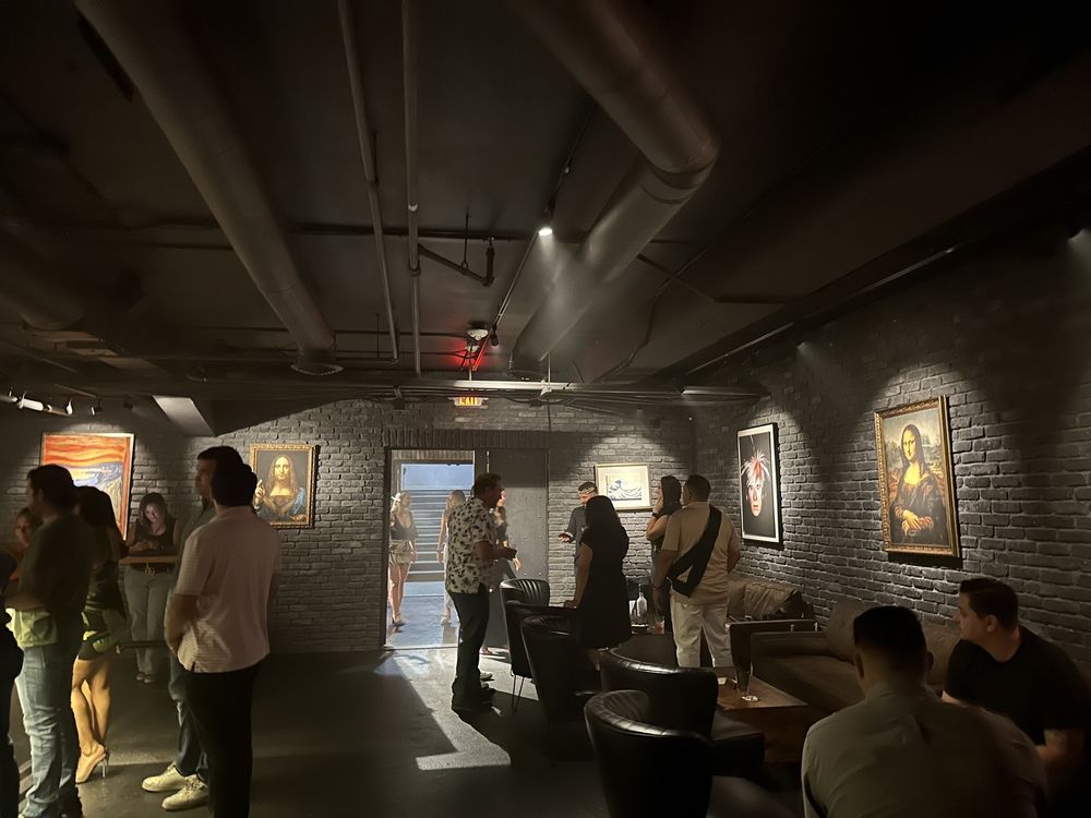 People in an art gallery with paintings on brick walls, some standing and some sitting, dim lighting, industrial ceiling.