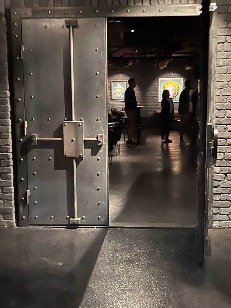 Open metal door leading into a room with people and stained glass windows, shadows cast on the floor.