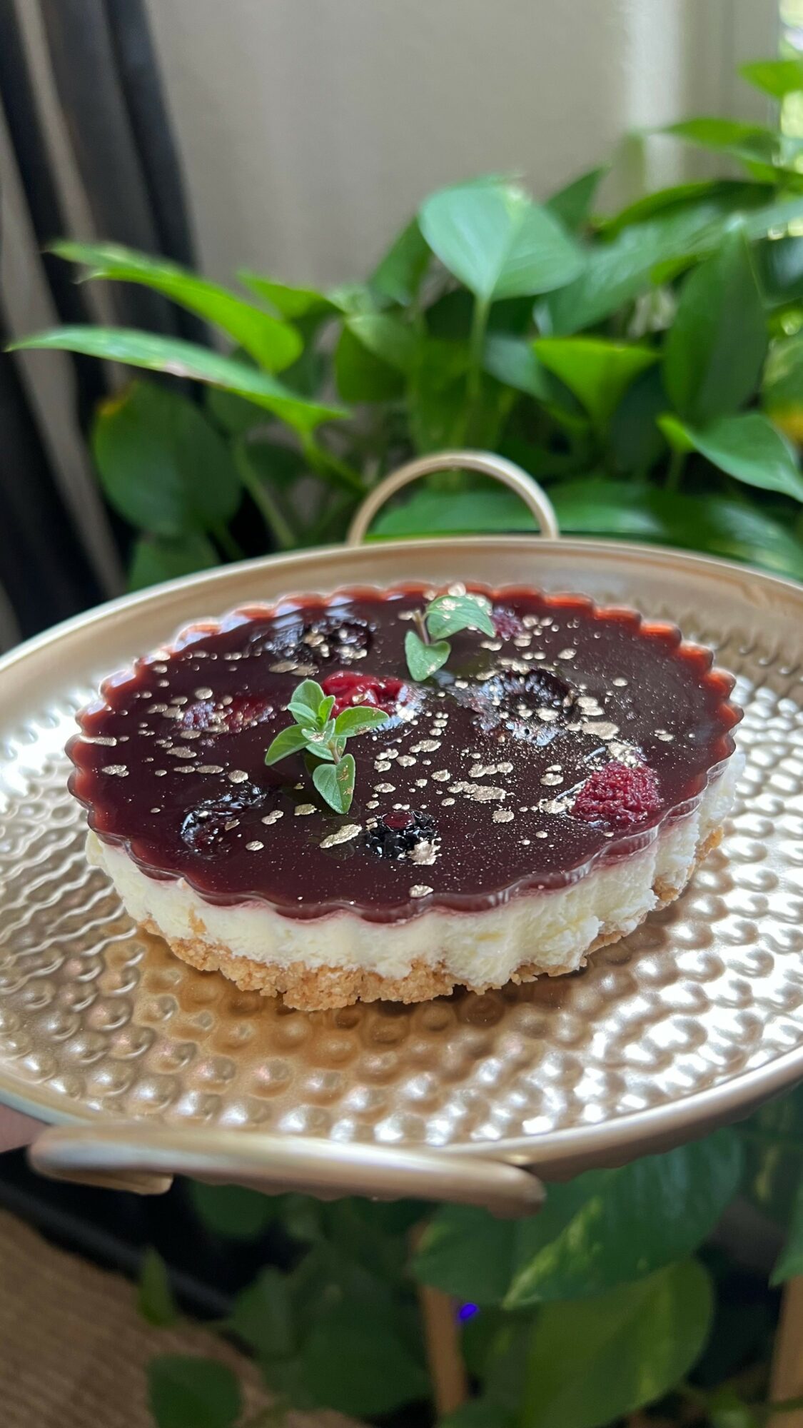 Cheesecake with berry topping, garnished with mint leaves, on a decorative plate near green plants.