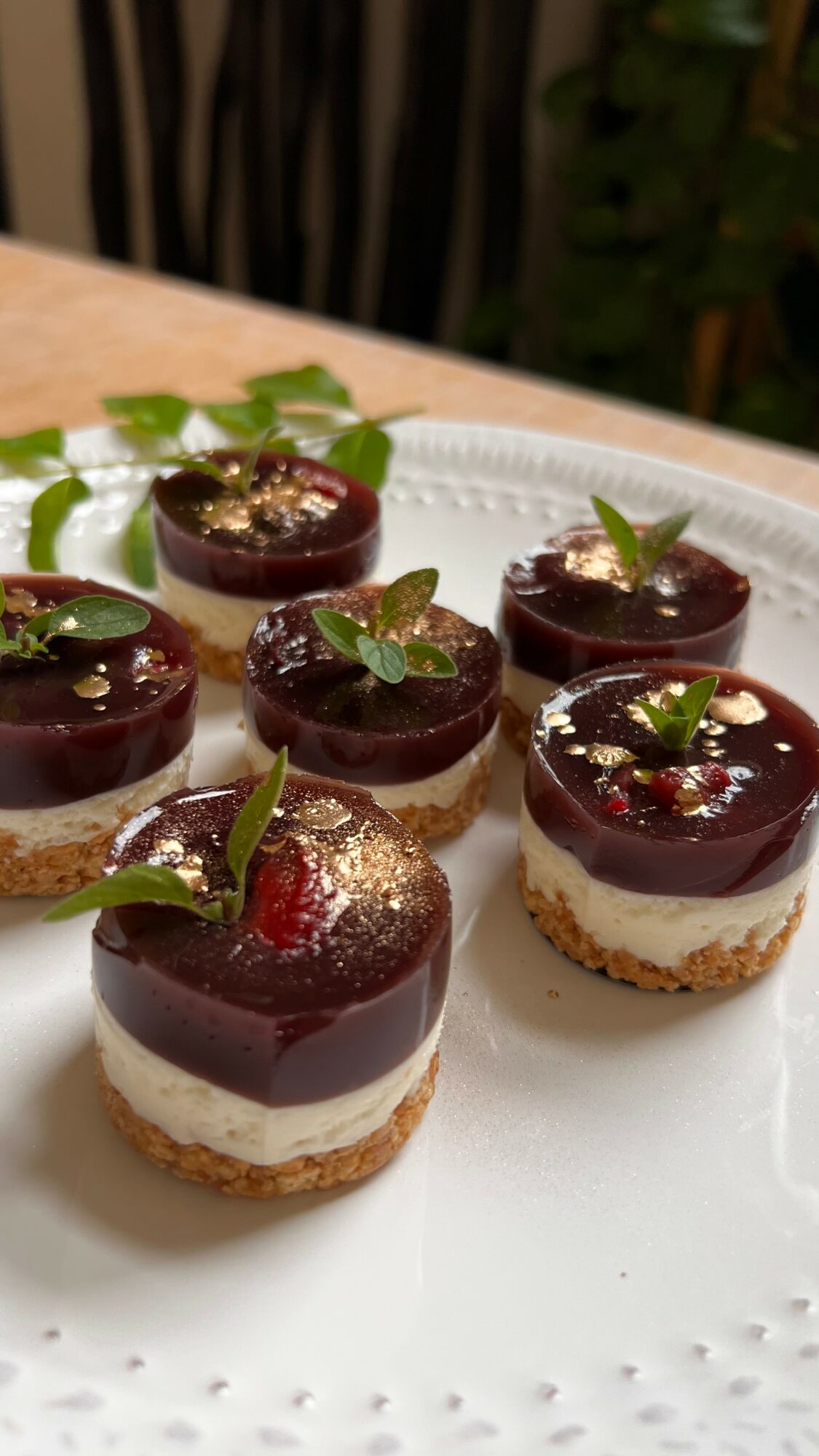 Six small layered desserts with dark glossy topping, garnished with mint leaves and edible gold, on a white plate.