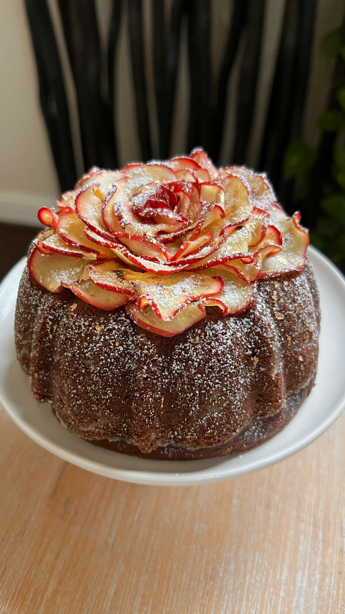 Chocolate cake topped with a decorative flower made of thinly sliced fruit, dusted with powdered sugar, on a white plate