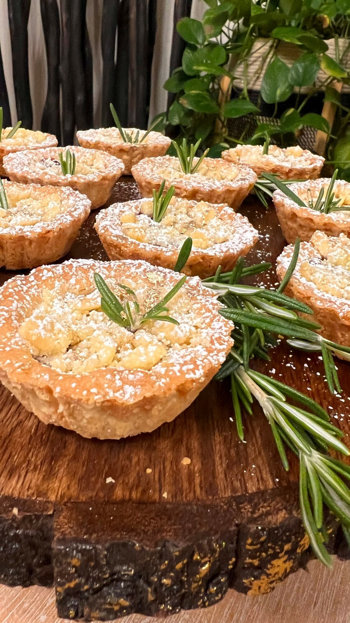 Multiple small tartlets topped with powdered sugar and garnished with green herbs, arranged on a wooden surface with greenery in the background.