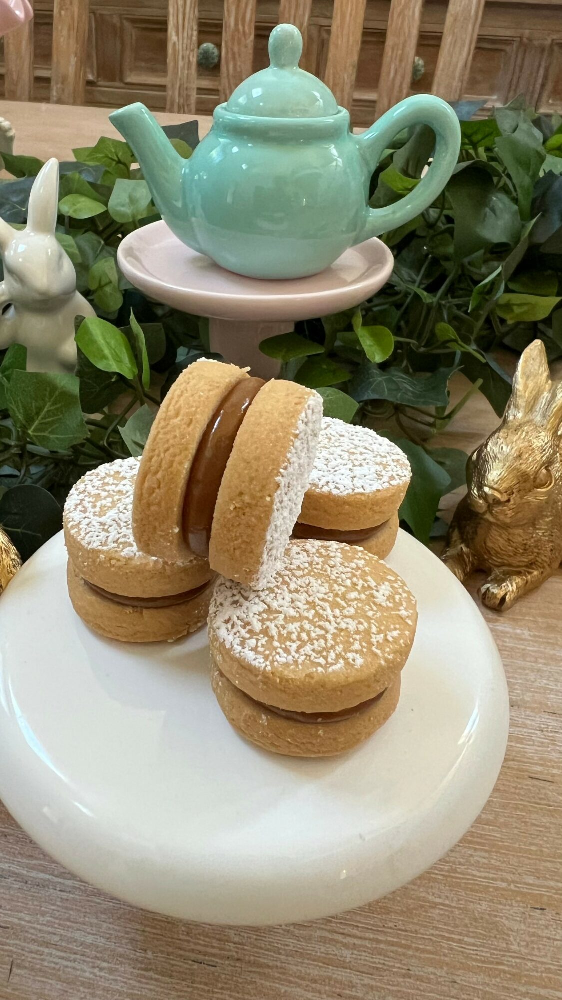 Plate with cookies, teapot and bunny figurine on a wooden table outdoors, surrounded by green plants and a wooden fence.