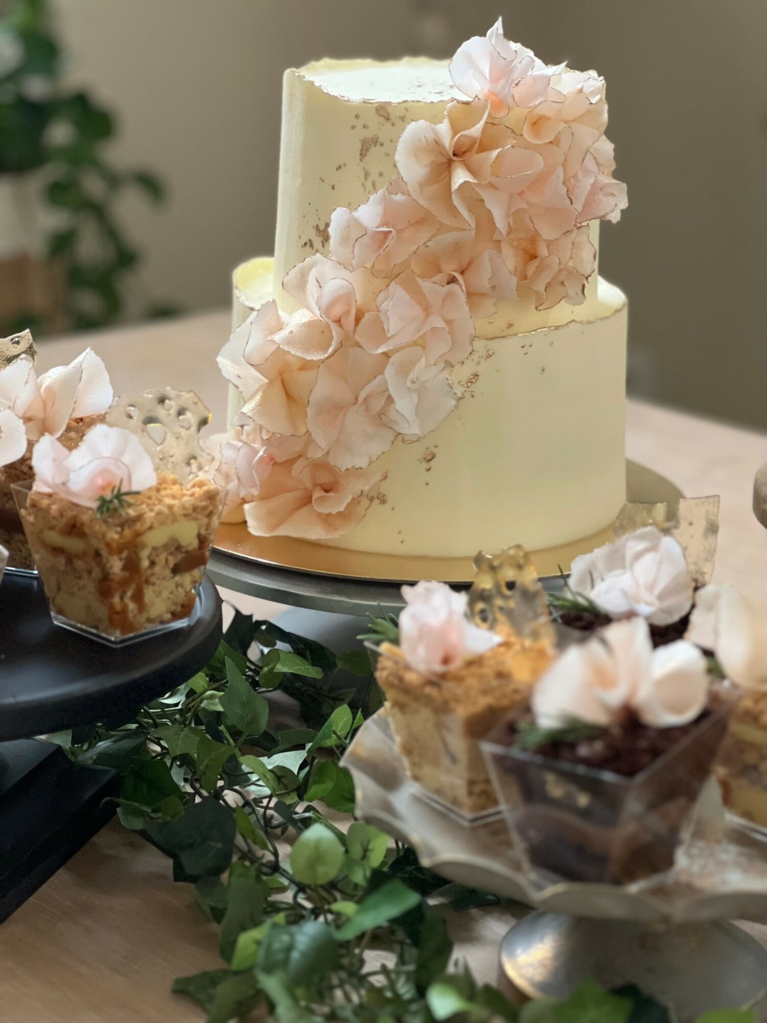 A two-tiered cake decorated with pink flowers, surrounded by small desserts on a table with greenery.