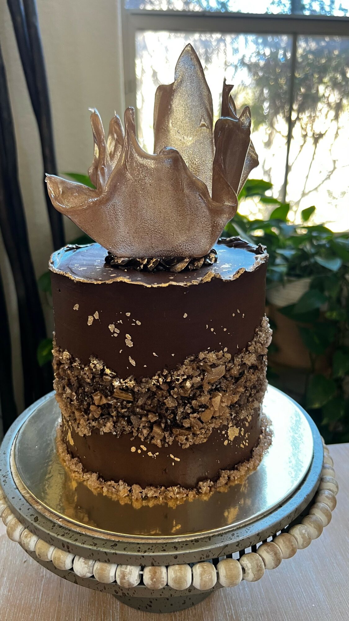 Chocolate cake with a metallic leaf decoration on top, placed on a decorative silver stand, with a window and greenery in the background.