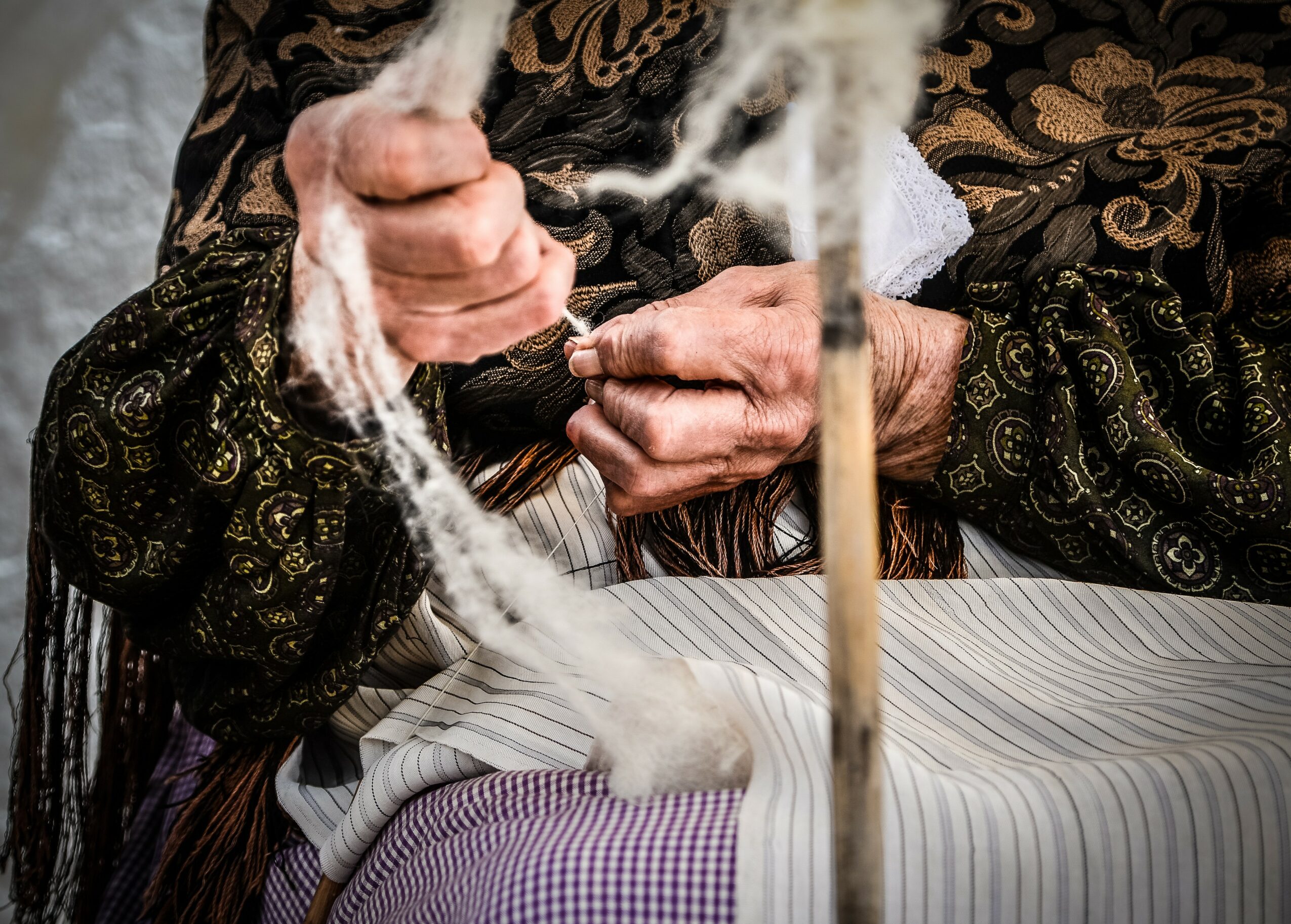 Person weaving white thread on a loom, wearing patterned clothing, with hands close-up, detailed fabric textures visible.