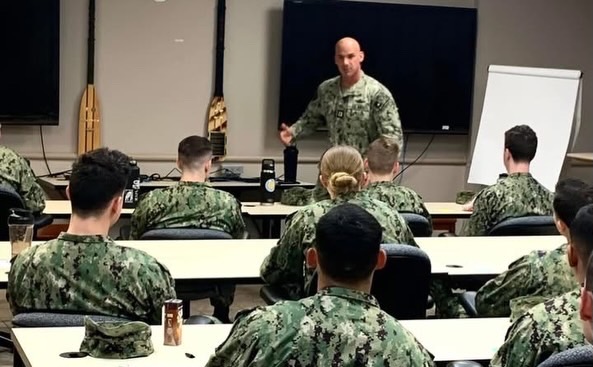 Military instructor standing in front of soldiers seated in classroom, with blackboard and whiteboard behind him.