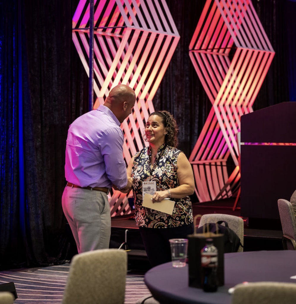 Two people, a man and a woman, are talking at an event with geometric pink and purple decorations in the background.