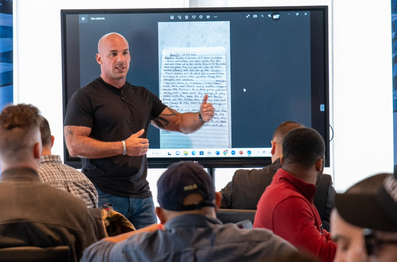 Man giving a presentation to a group in a classroom or conference room, with a large screen behind him displaying a document.