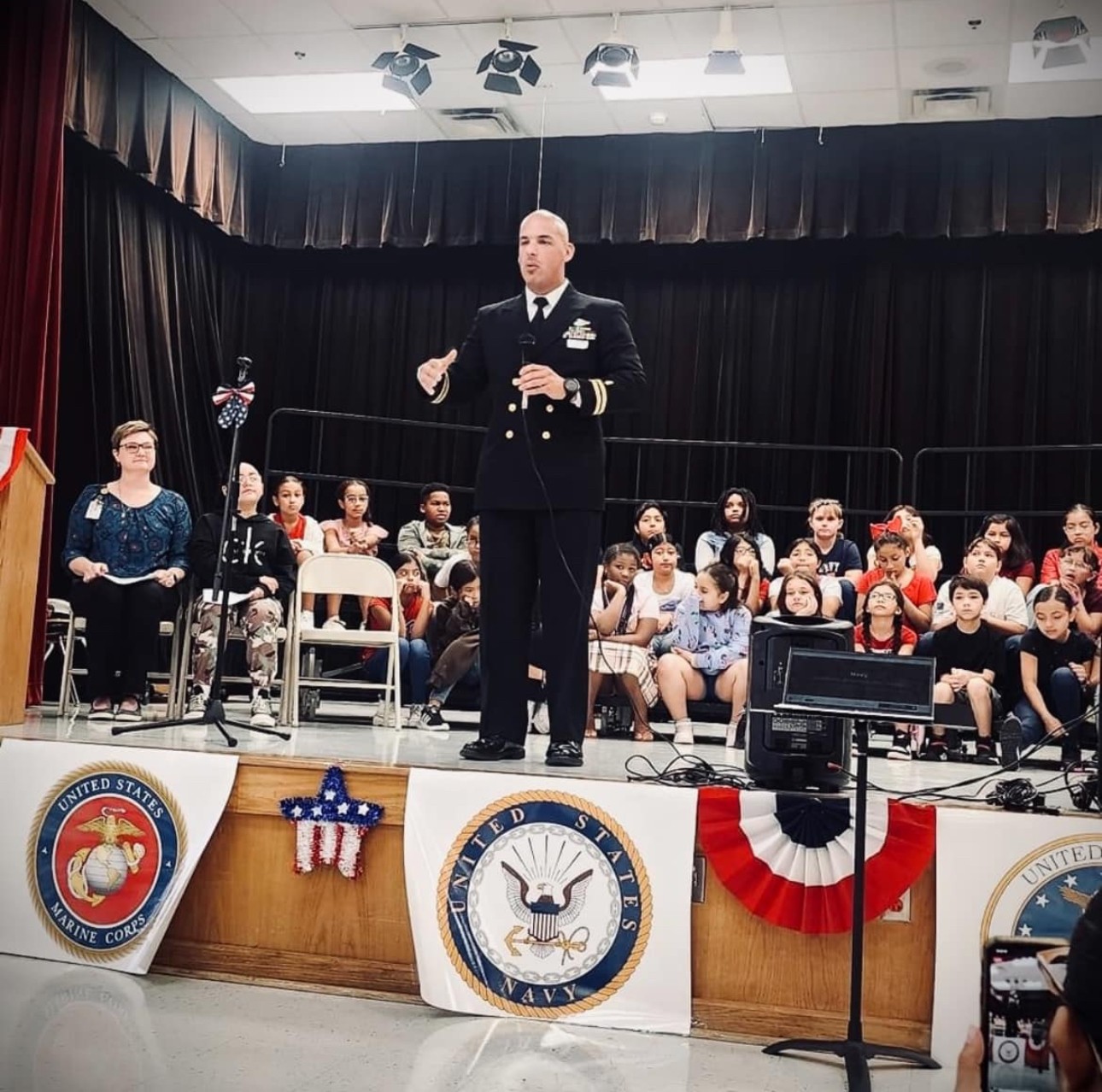 Man in military uniform speaking on stage with children and adults seated behind him, decorated with patriotic banners.