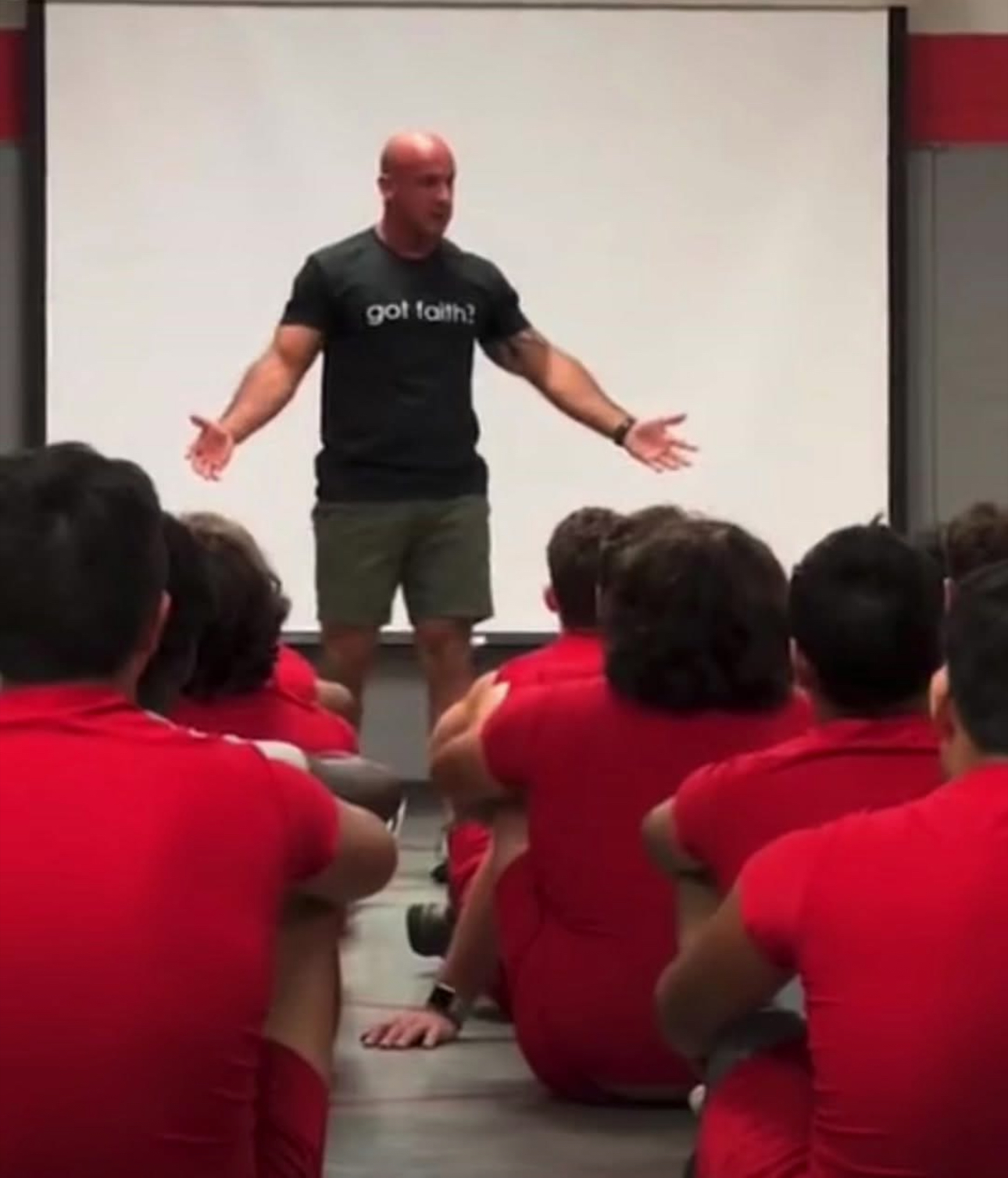 A man stands in front of a group of people wearing red shirts, gesturing with arms open, in a room with a white screen behind him.
