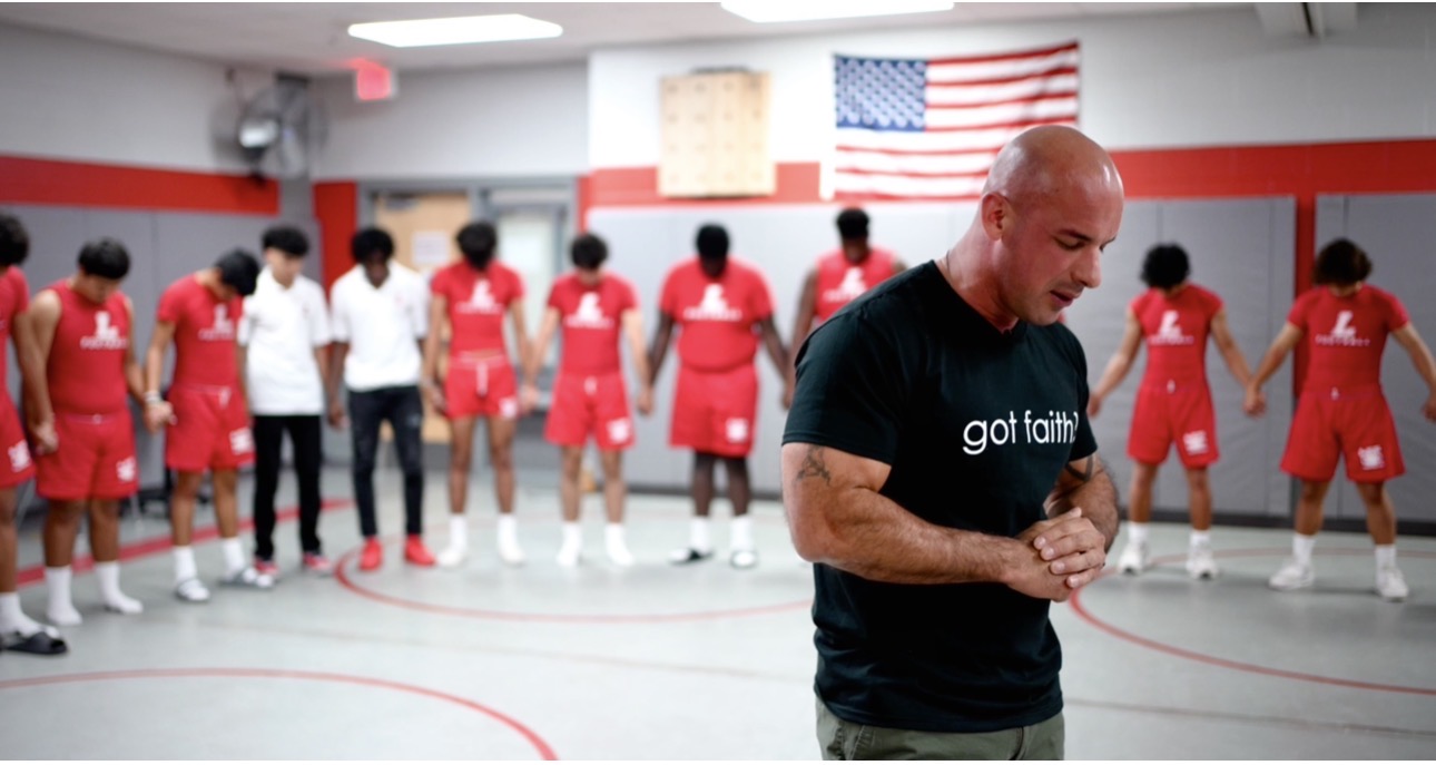 Basketball team in red uniforms lined up on court with coach praying in foreground, American flag on wall behind.