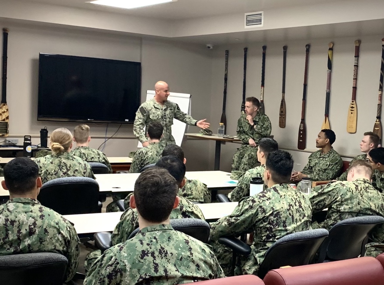 Military personnel in camouflage uniforms attending a classroom session with two officers at the front, one speaking and the other listening, rifles on the wall behind them.