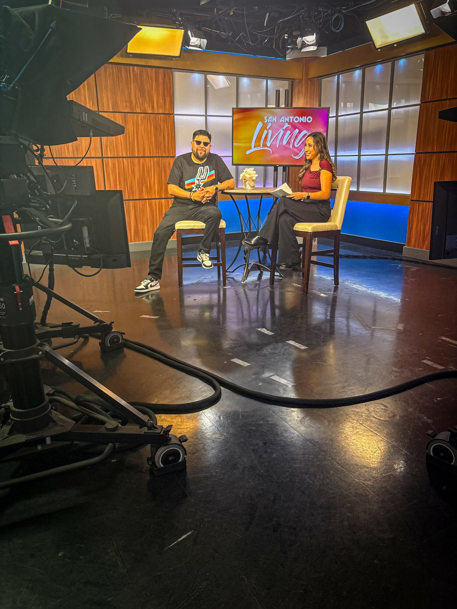 Two people sit at a table on a TV set with a wooden background and a screen displaying 'Vive' behind them.