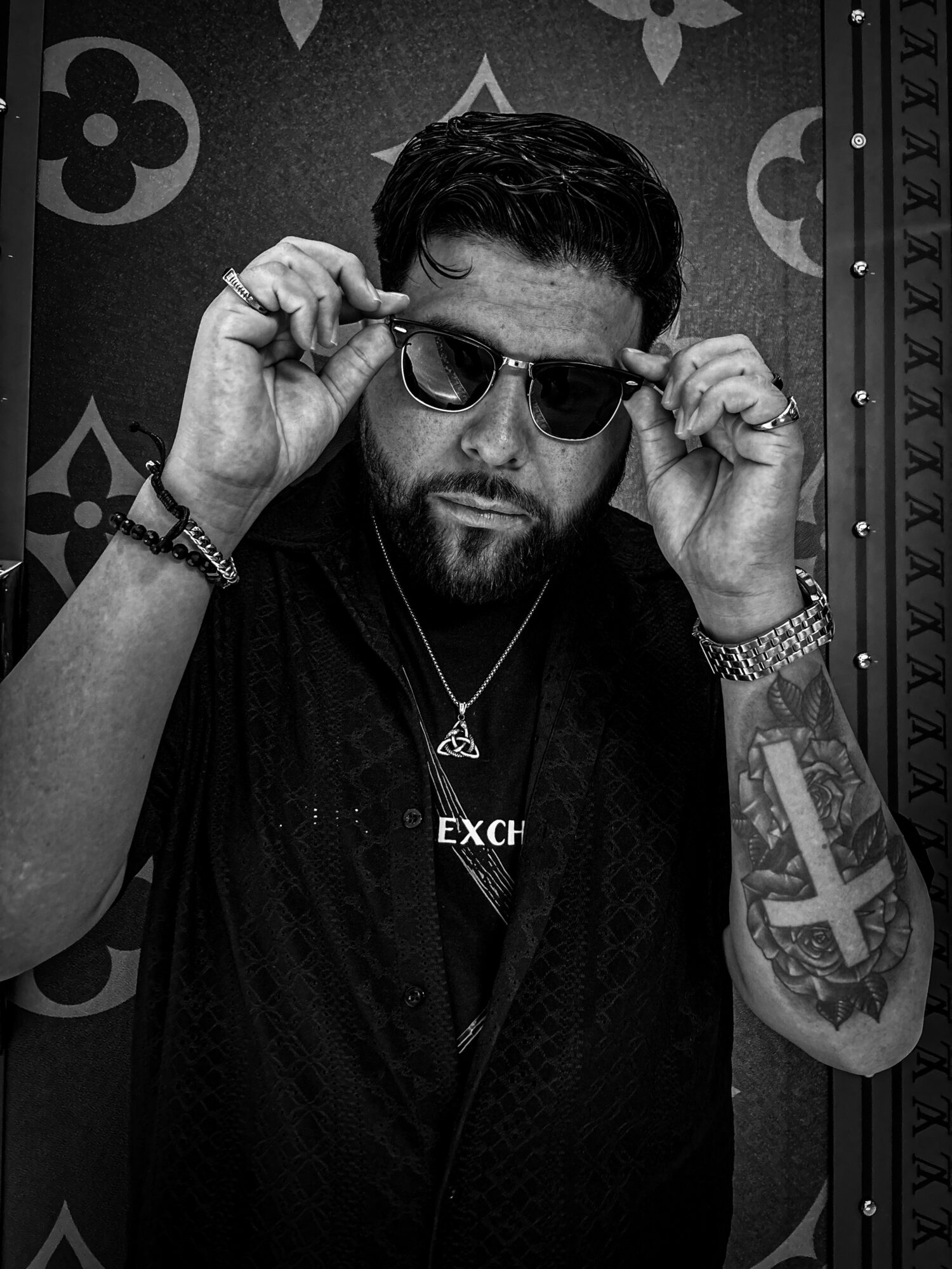 Man with sunglasses adjusting them, wearing a necklace and watch, with a tattoo on his arm, in front of patterned background.